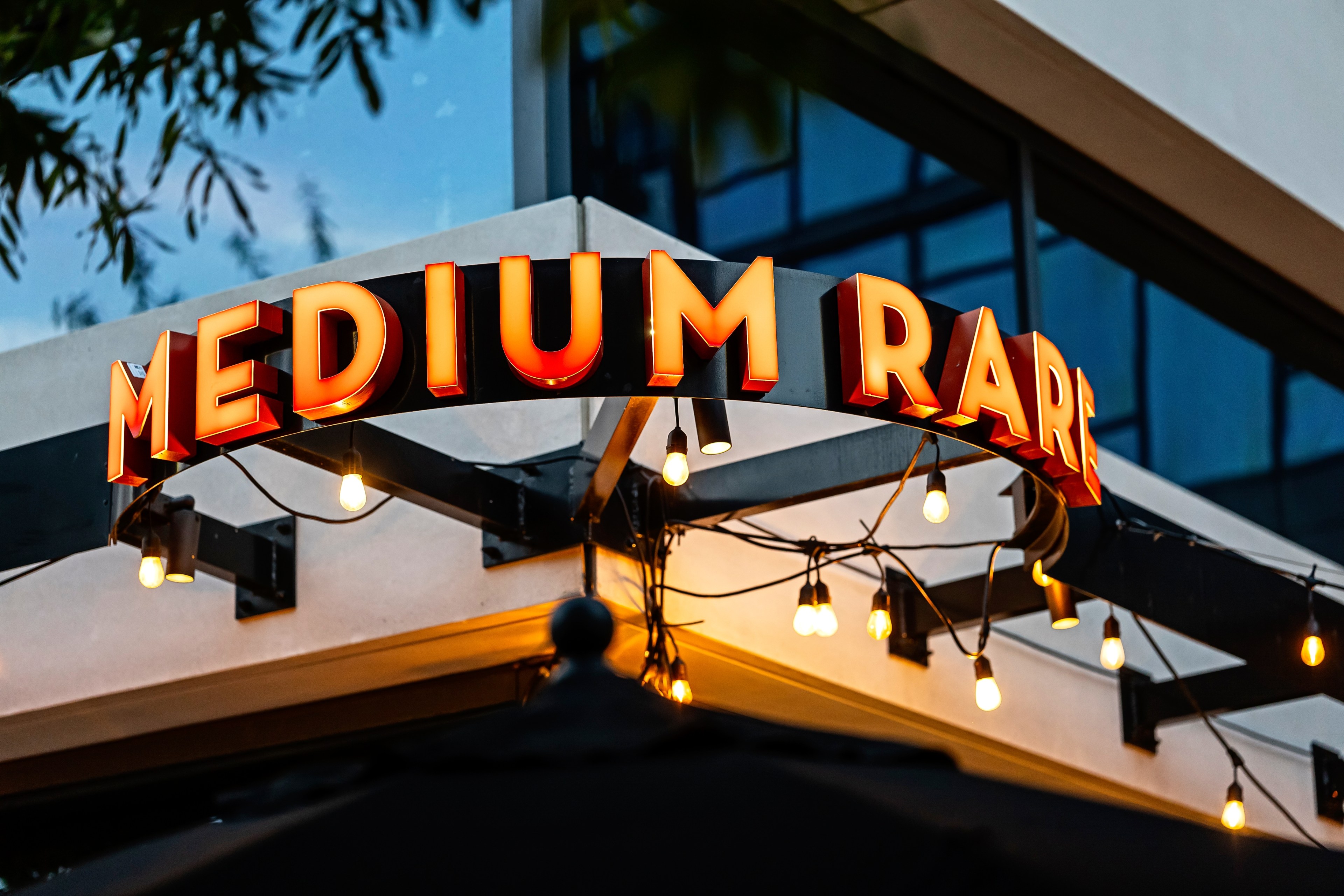 Glowing orange letters spell out the words "Medium" and "Rare" against a circular metal frame above a restaurant entrance.