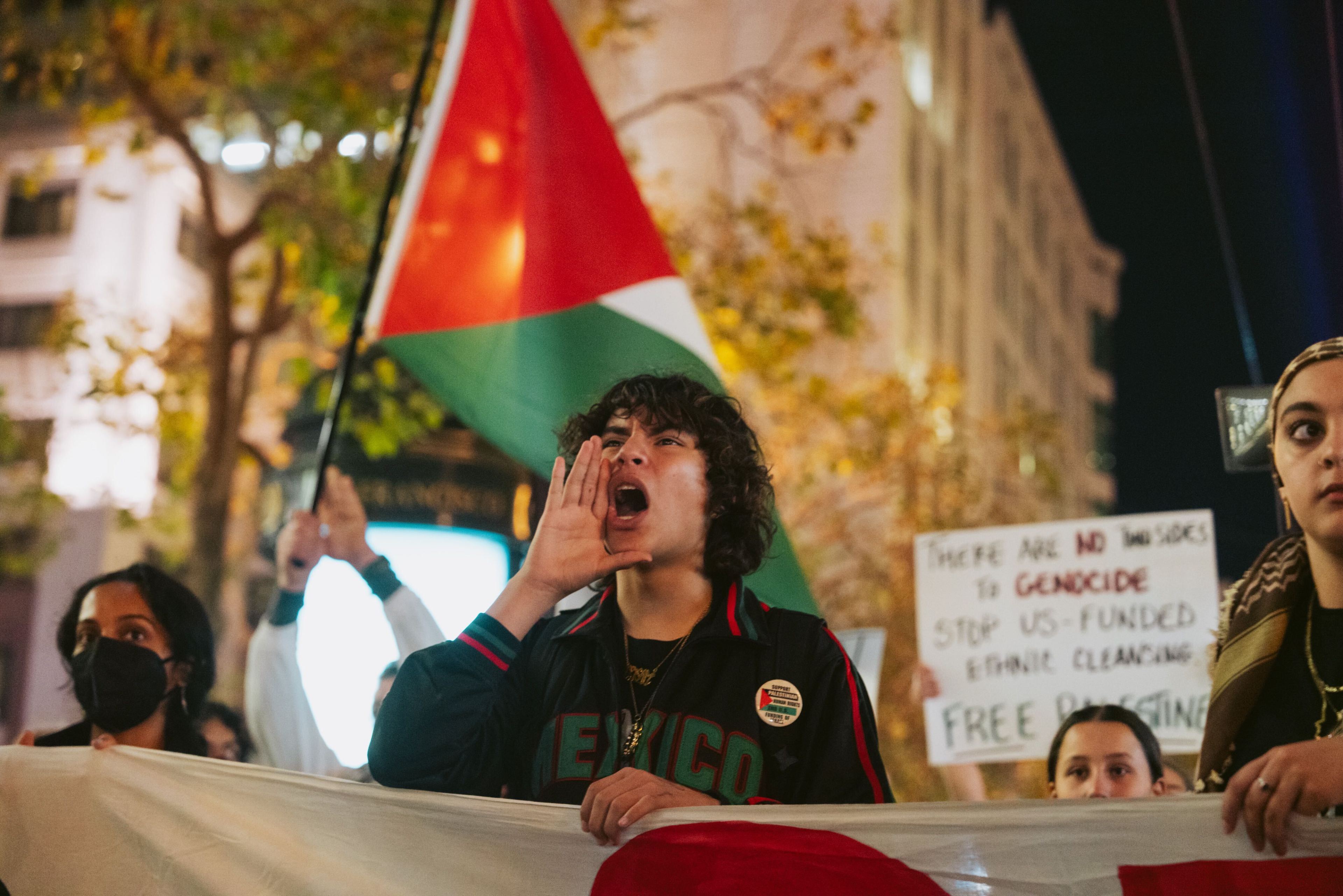 A person chanting at a protest while holding a banner and marching beside a Palestinian flag.