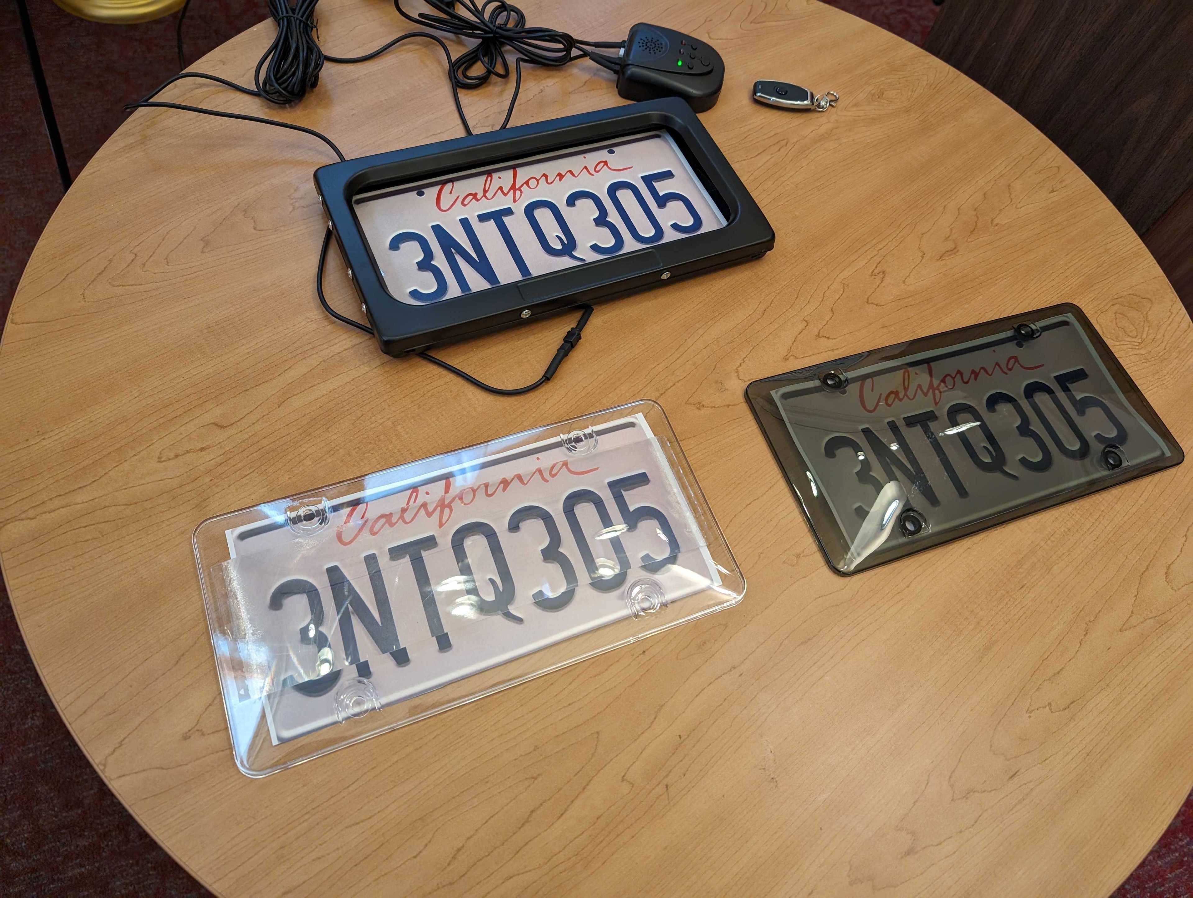 A circular wooden tabletop holds three versions of the same California license plate. One version is in an automatic device that responds to a key fob button, shuttering a sliding black door over the plate. Another version covers the plate with semitransparent vinyl that distorts the letters and numbers, while a third version tints and darkens the plate to limit its legibility.