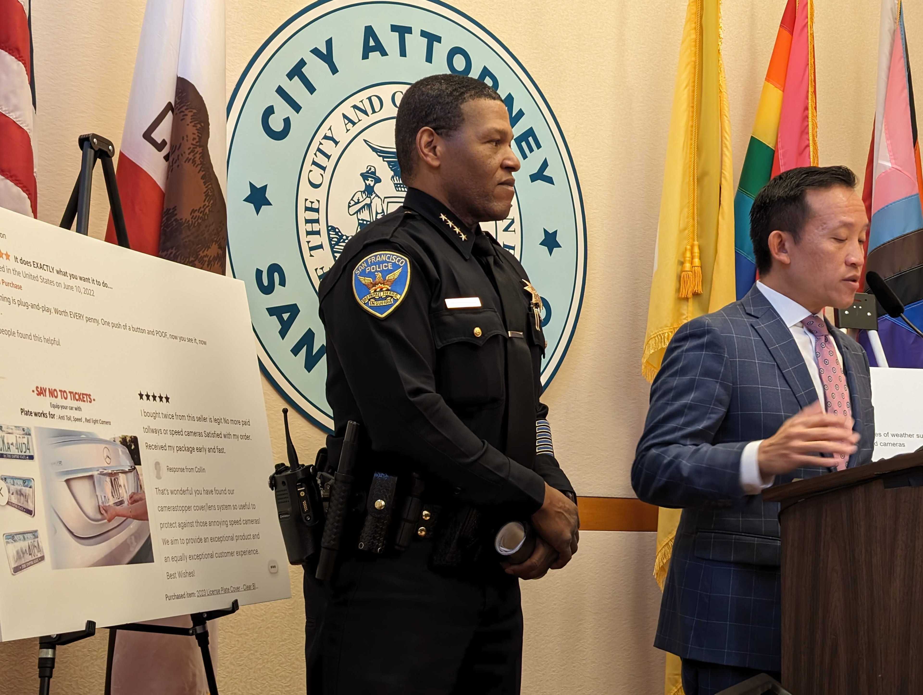 A uniformed police chief stands beside a placard showing images and online reviews of illegal license plate holders. The chief actively listens to a suit-wearing man standing beside him at a podium and speaking at a press conference. A San Francisco City Attorney seal and American, state, city and pride flags stand on a wall behind the two men.