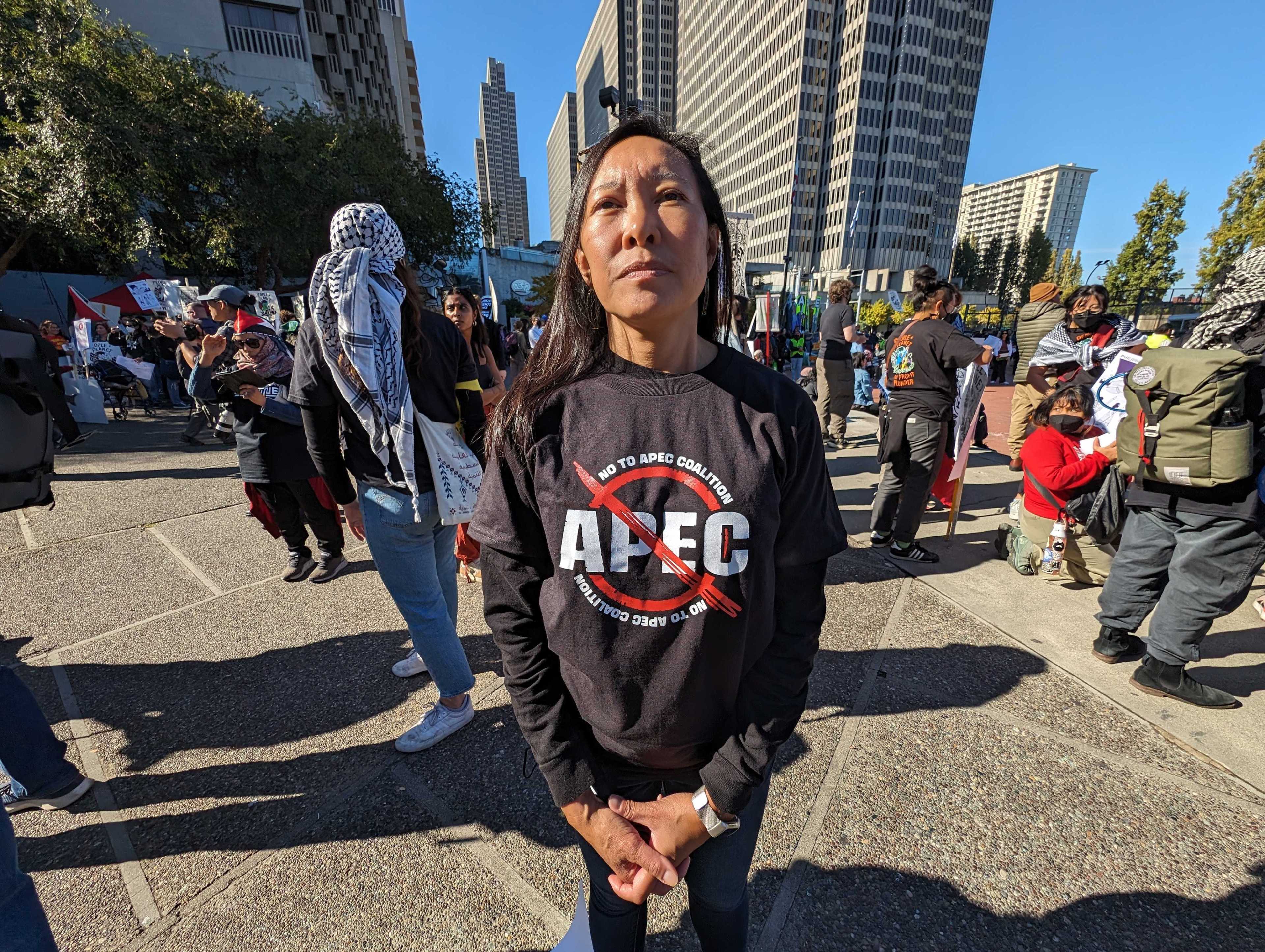 A woman wears a black &quot;No to APEC Coalition shirt&quot; while standing in a crowd.