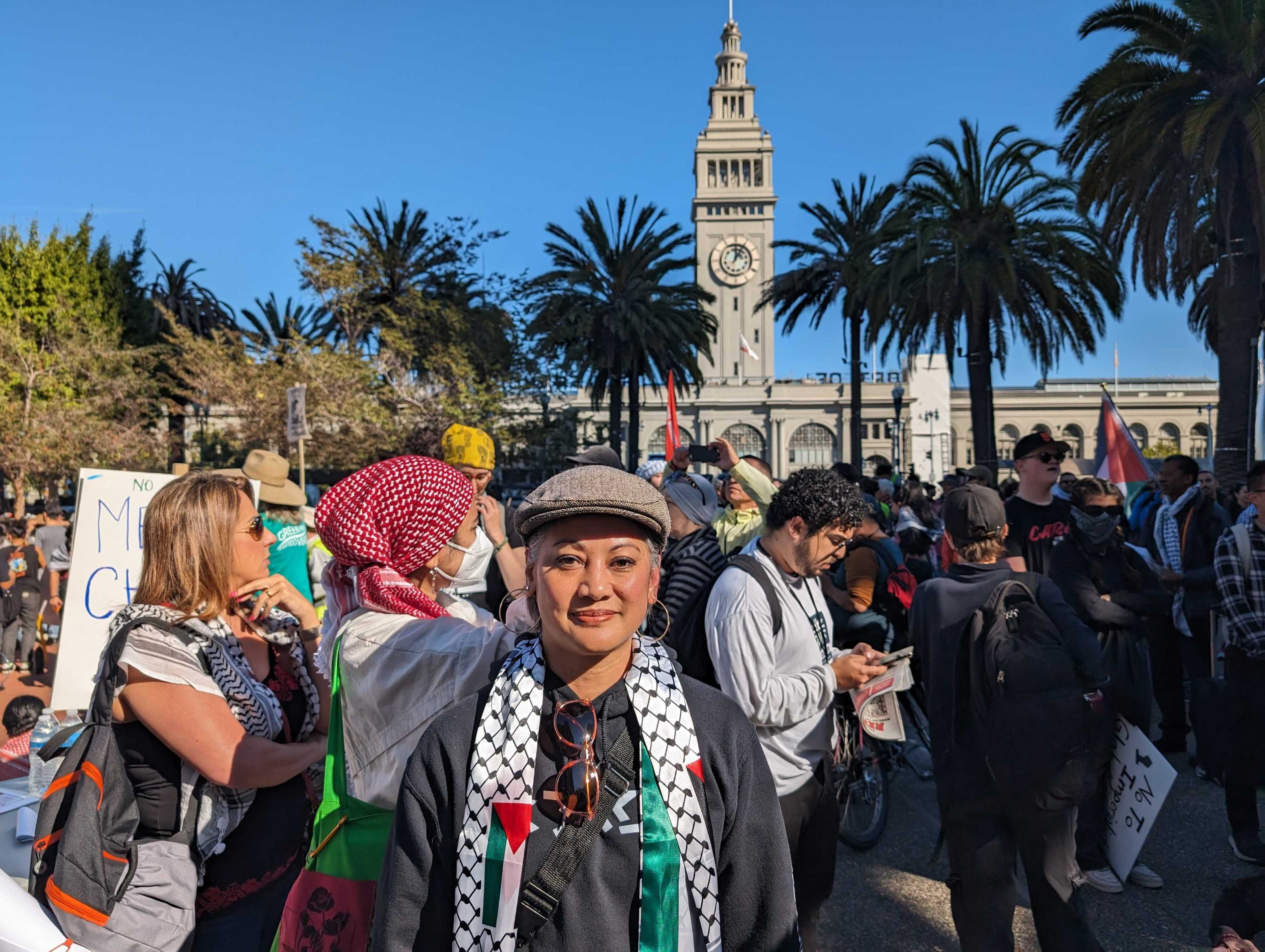 A woman in a hat and scarf smiles alongside protesters outside under blue skies and palm trees. 