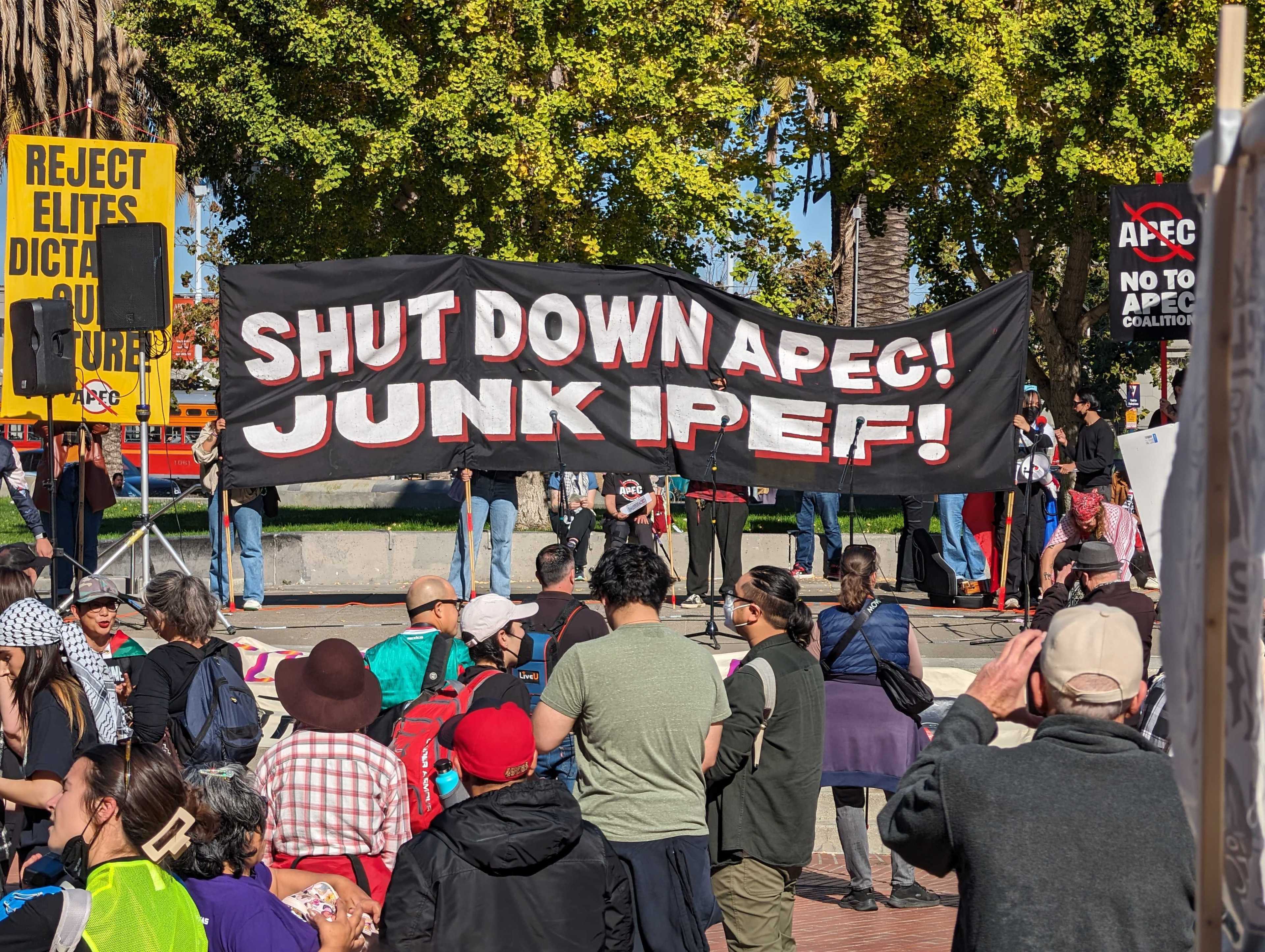 A black banner that says Shut Down APEC! Junk IPEF! is displayed in a public plaza