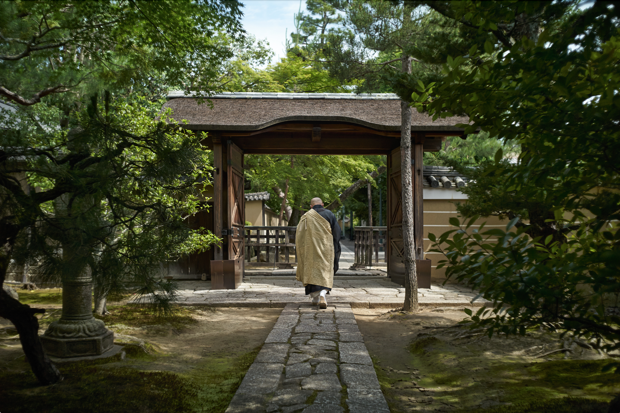 A robed man walks through a gate that has trees and greenery all around it.