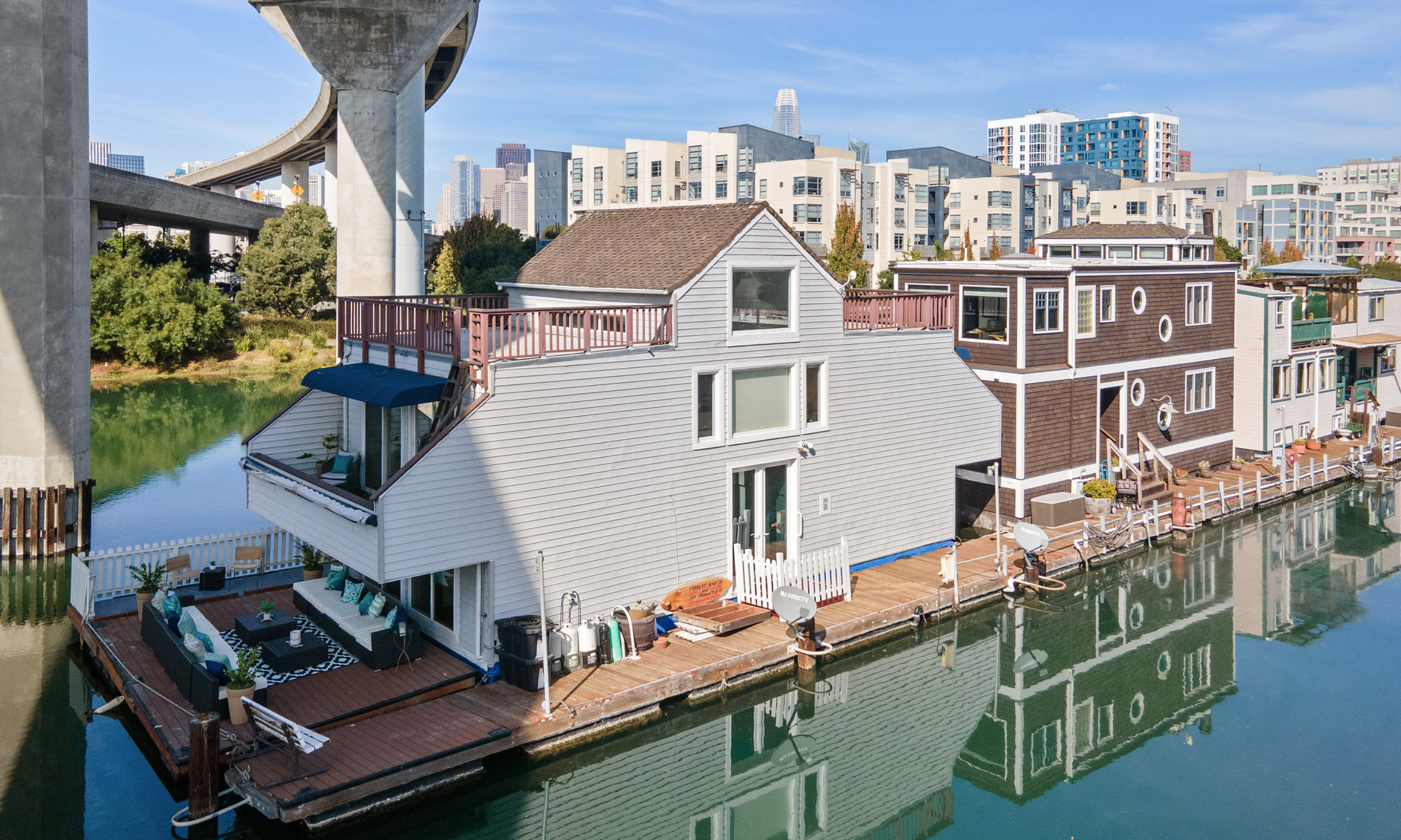 A triangular, gray houseboat sits at the end of a dock in Mission Creek on a sunny day.