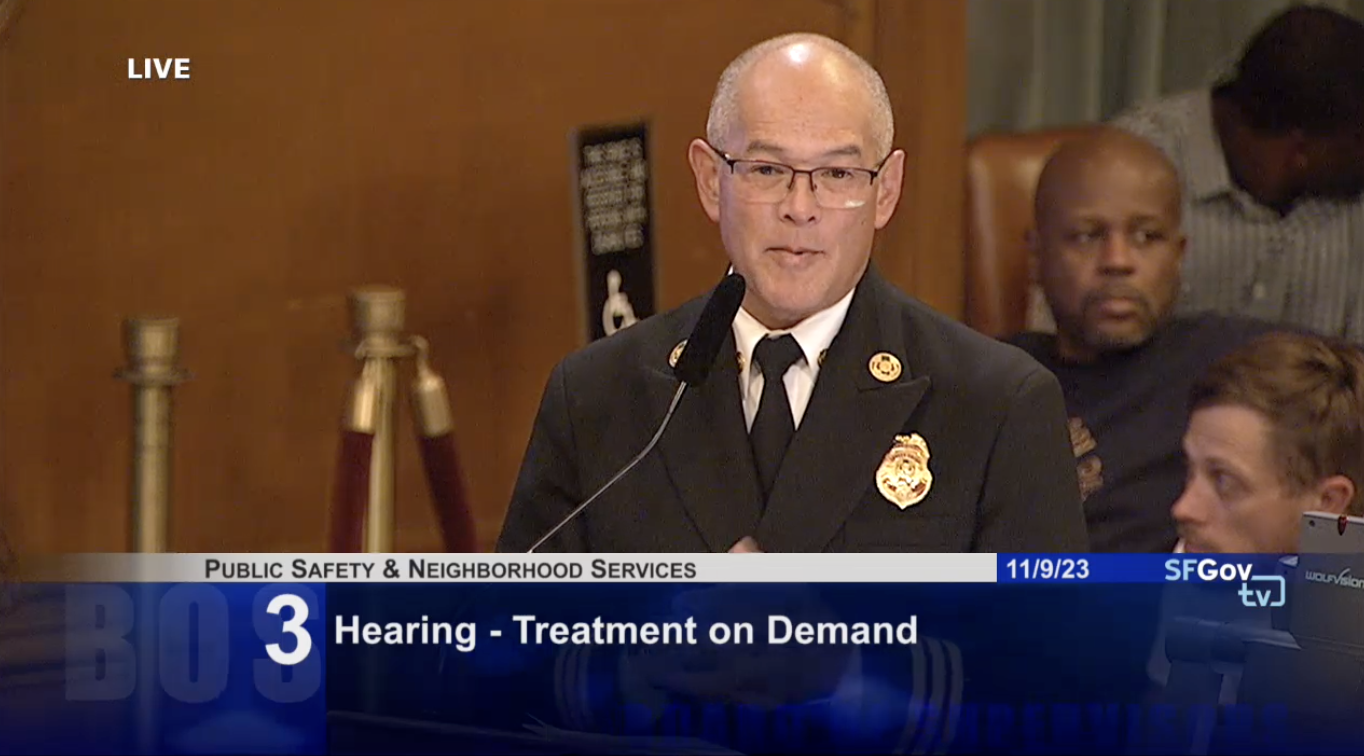 A man wearing a shirt, tie and blazer with a San Francisco Fire Department badge is seen speaking at a podium from the torso up.