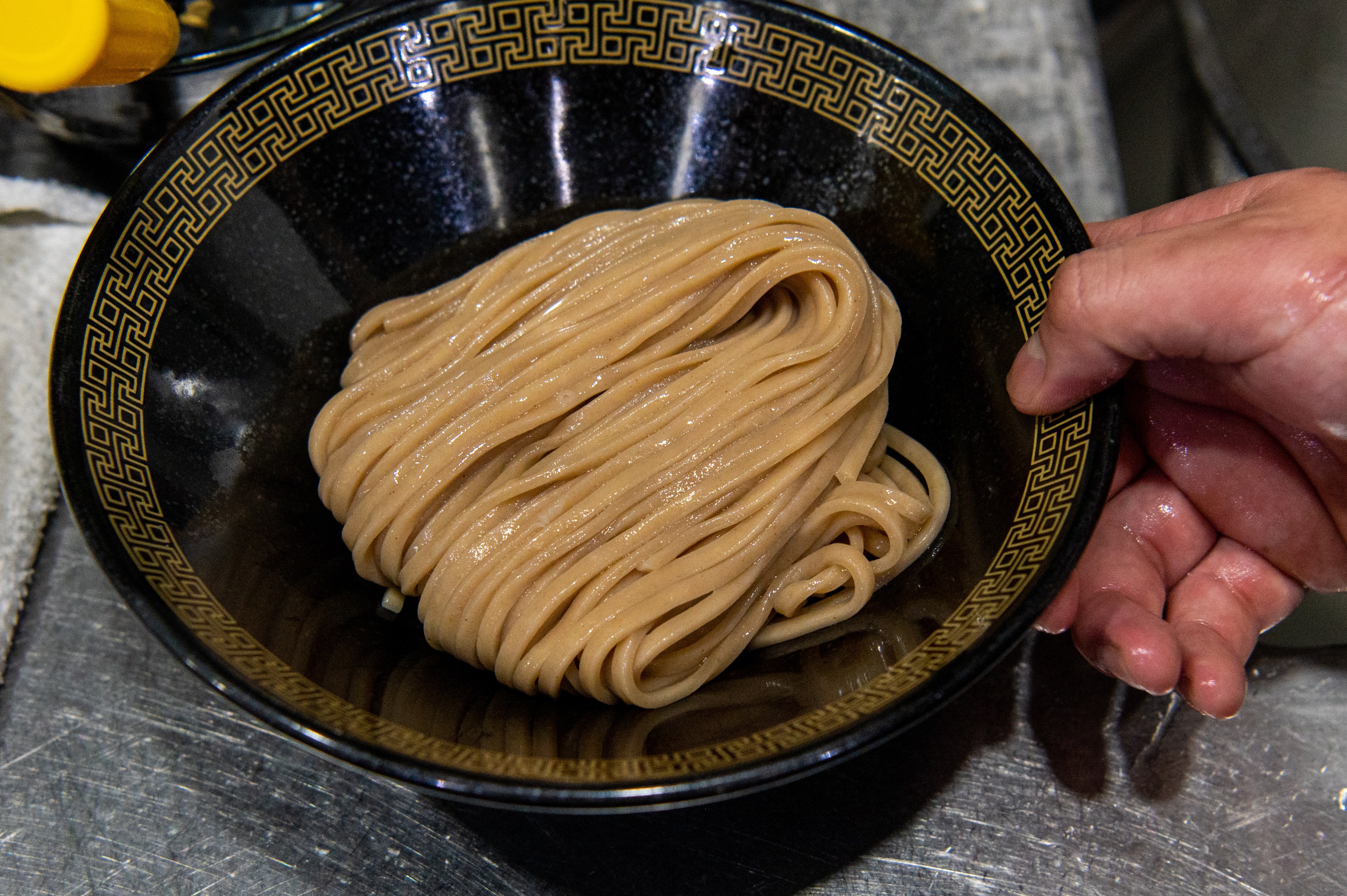 A hand holds a black bowl with gold trim filled with noodles.