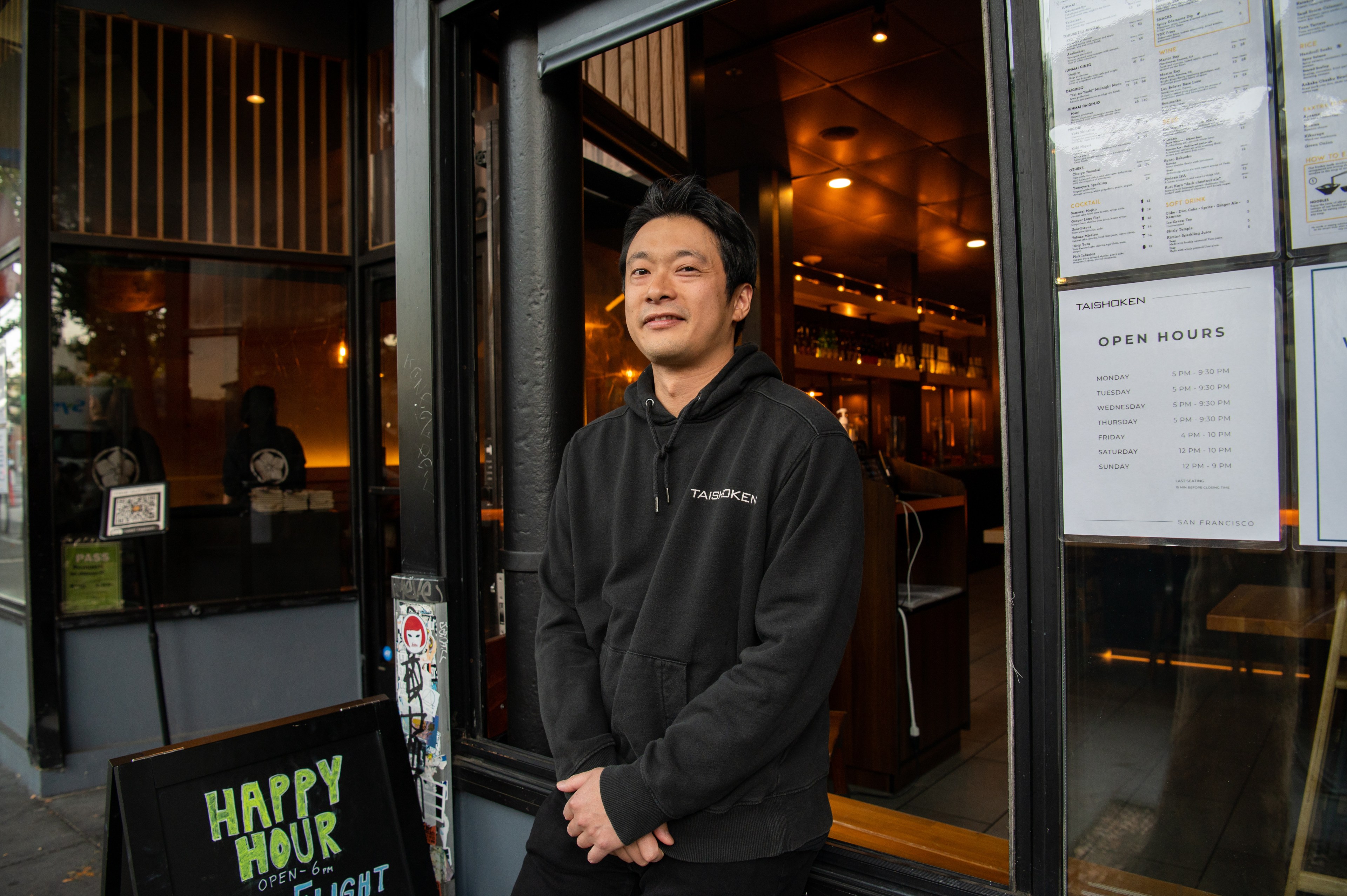 A man in a black hoodie stands with his hands clasped in front of him while standing in front of a restaurant's glass doors.