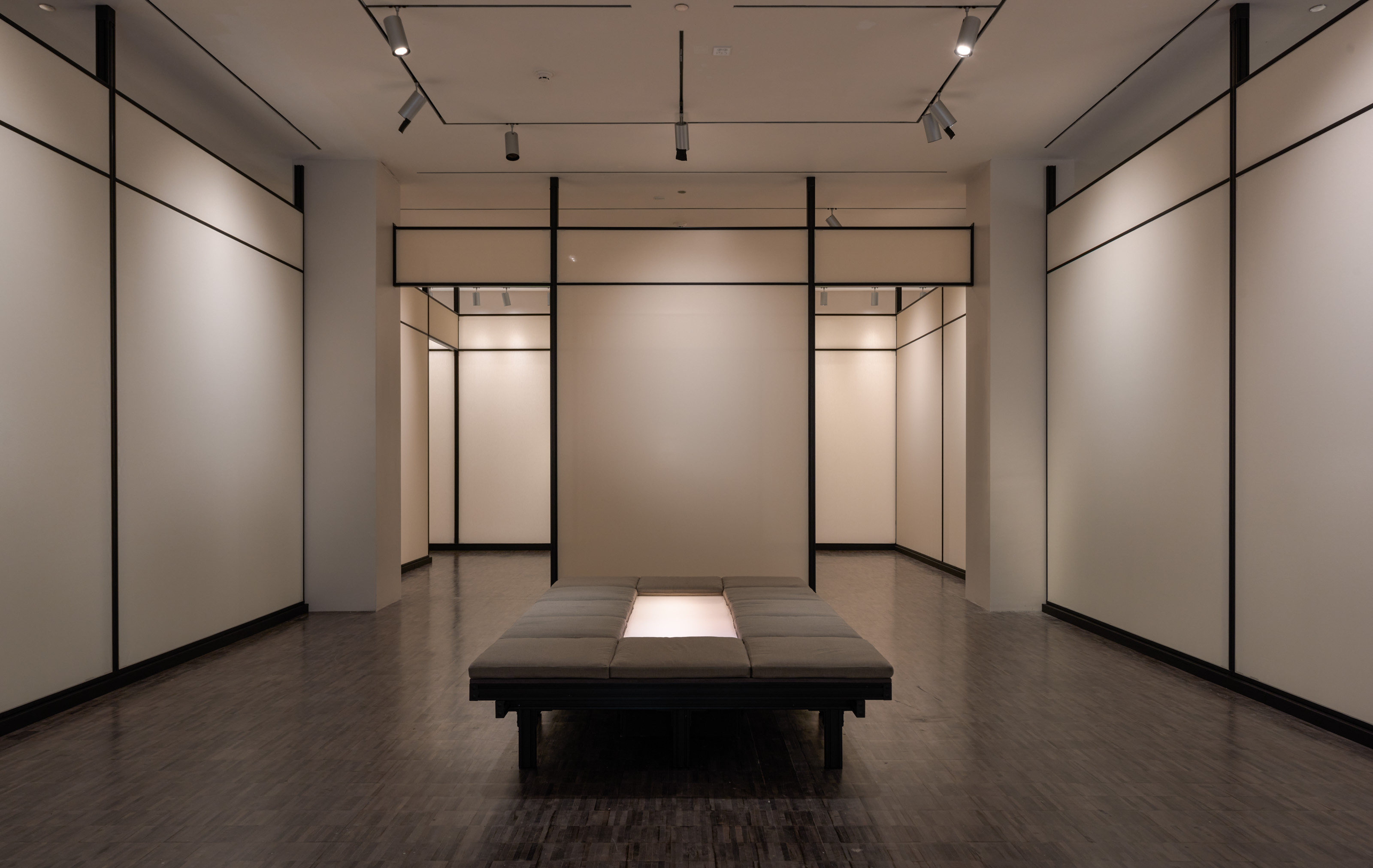 A view of a gallery space that has beige walls edged with black and a low, taupe-colored bench in the middle. 