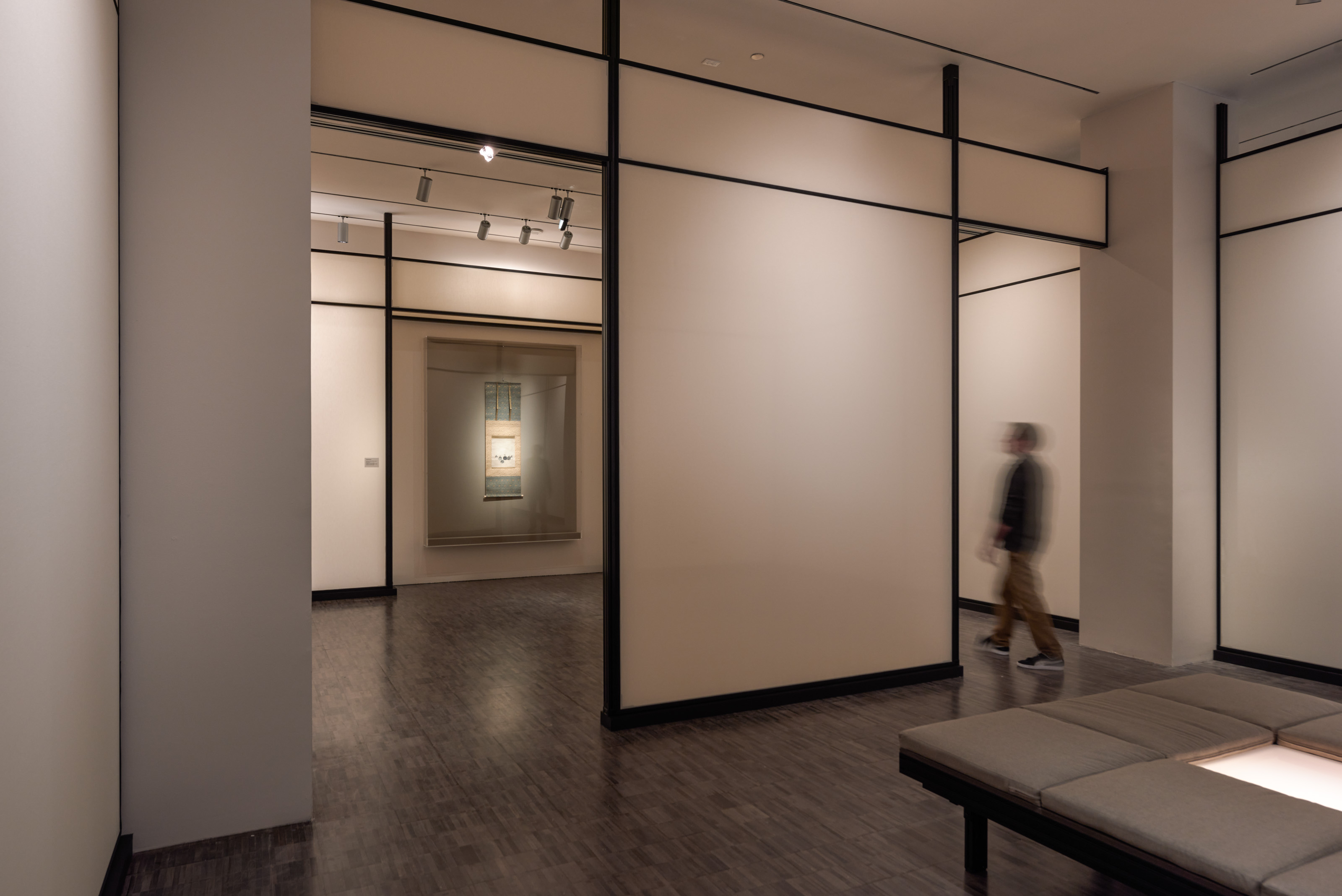 A person walks through a beige-colored gallery that has a piece of art in the distance hanging on the wall.