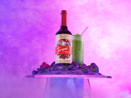 A bottle labeled Torani stands against a purple background with a green-colored beverage standing next to it.