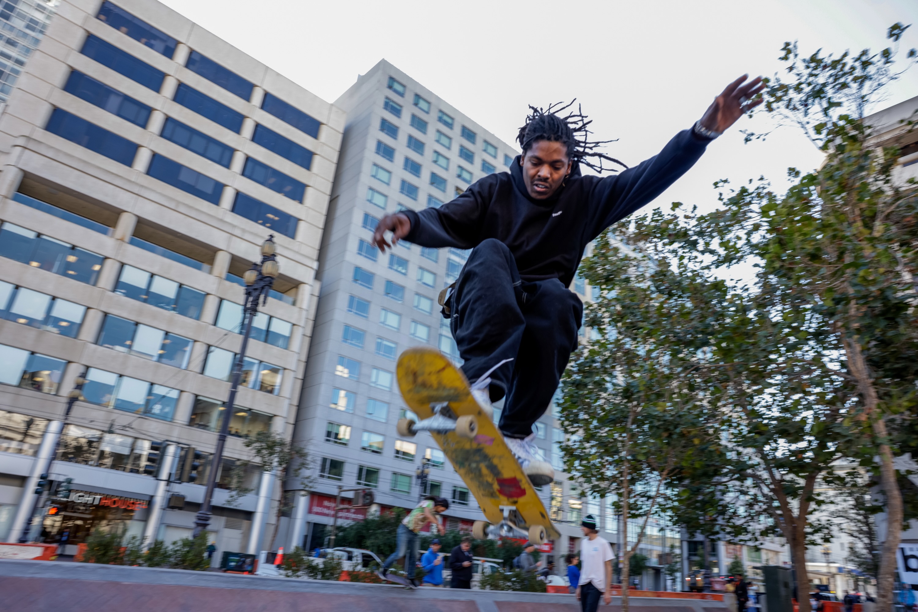 A person on a skateboard does a trick off a jump on a red brick skate park at UN Plaza with large glass buildings in the background in downtown San Francisco.