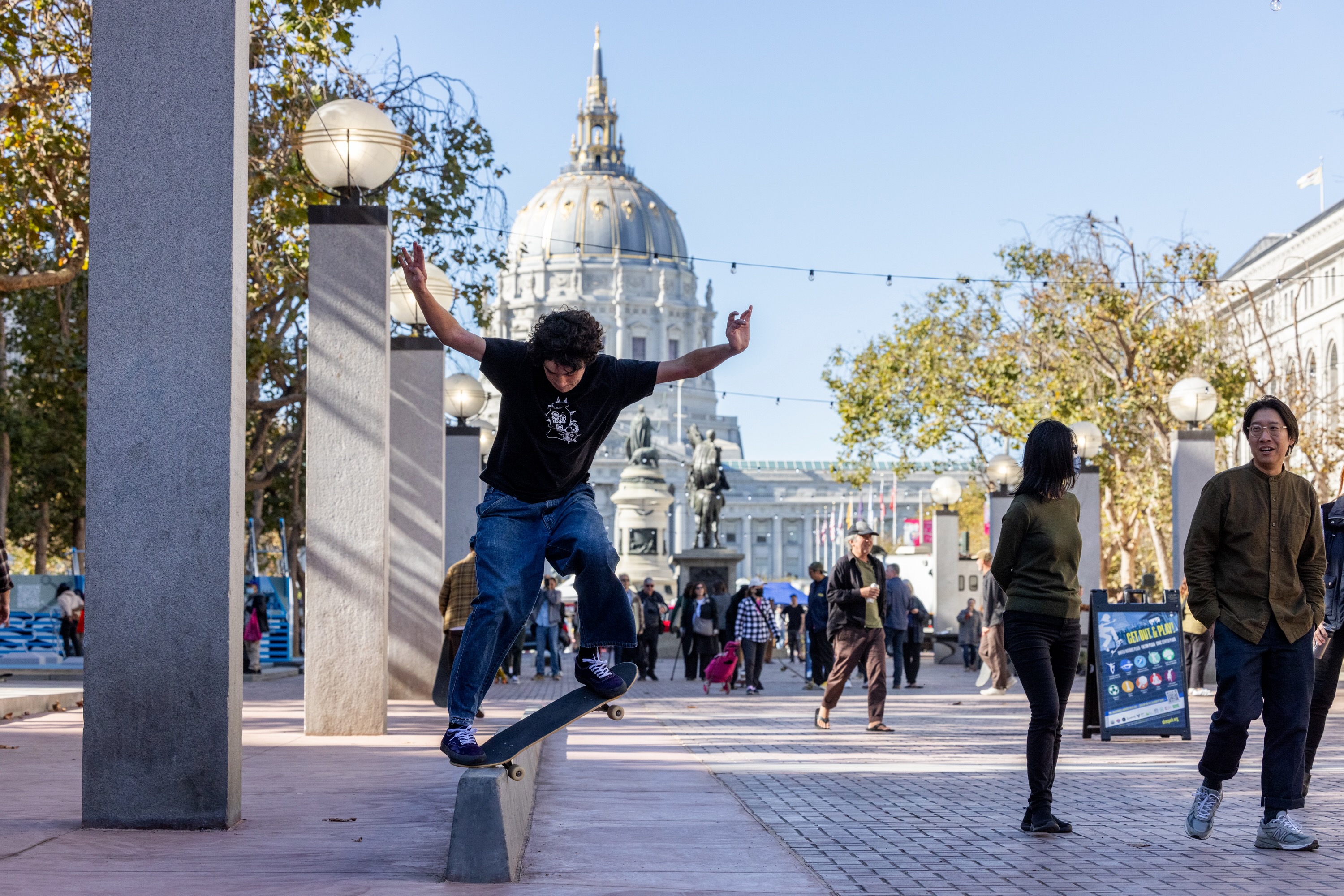 A person on a skateboard does a trick off a jump on a red brick skate park at UN Plaza with City Hall in the background on a sunny day in downtown San Francisco.