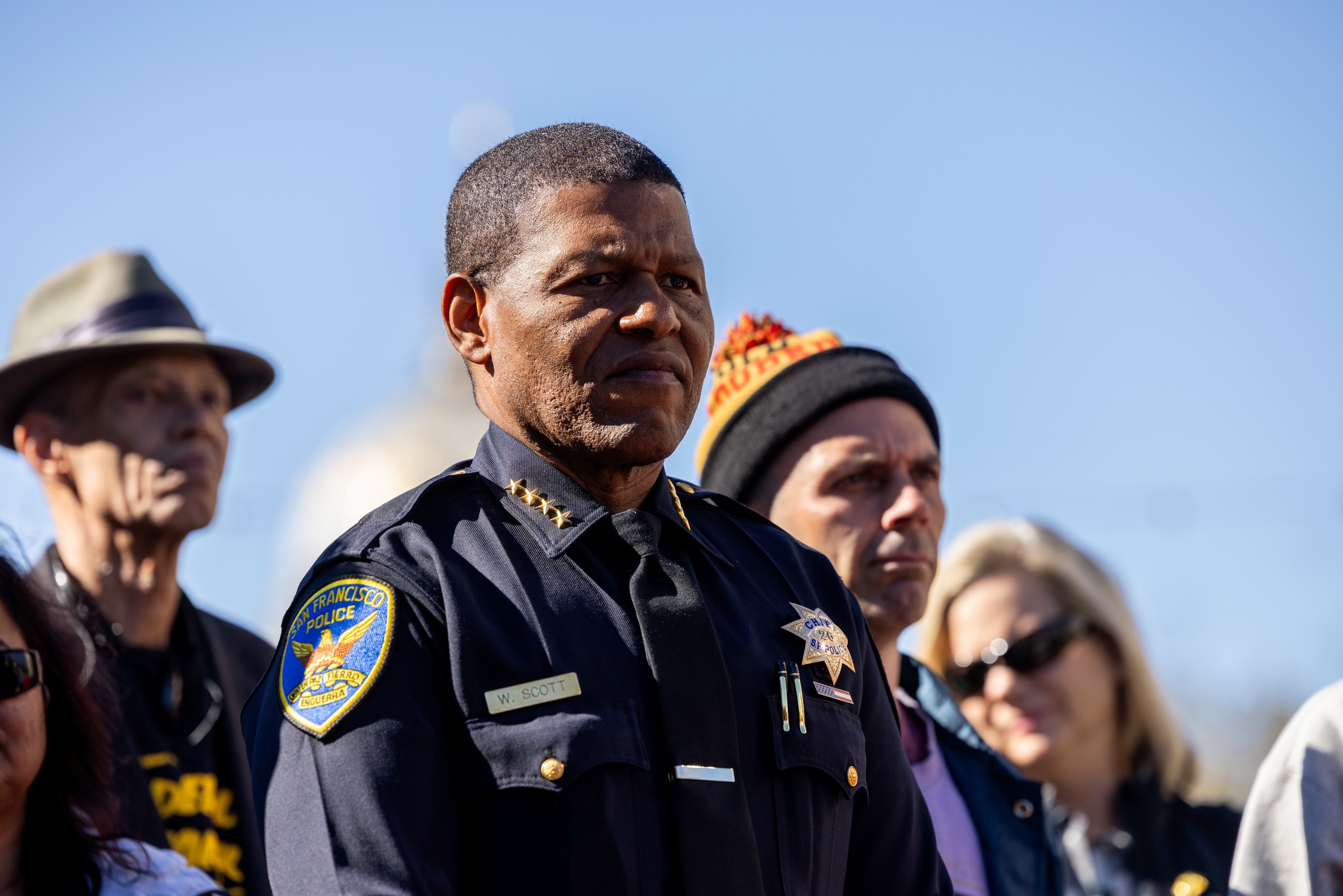 San Francisco Police Department Police Chief Bill Scott, wearing a police uniform looks into the distance with civilians standing behind him. Scott joined Mayor London Breed, elected officials, Tenderloin residents, and community partners to celebrate the reopening of the U.N Plaza which included a skateboard park.