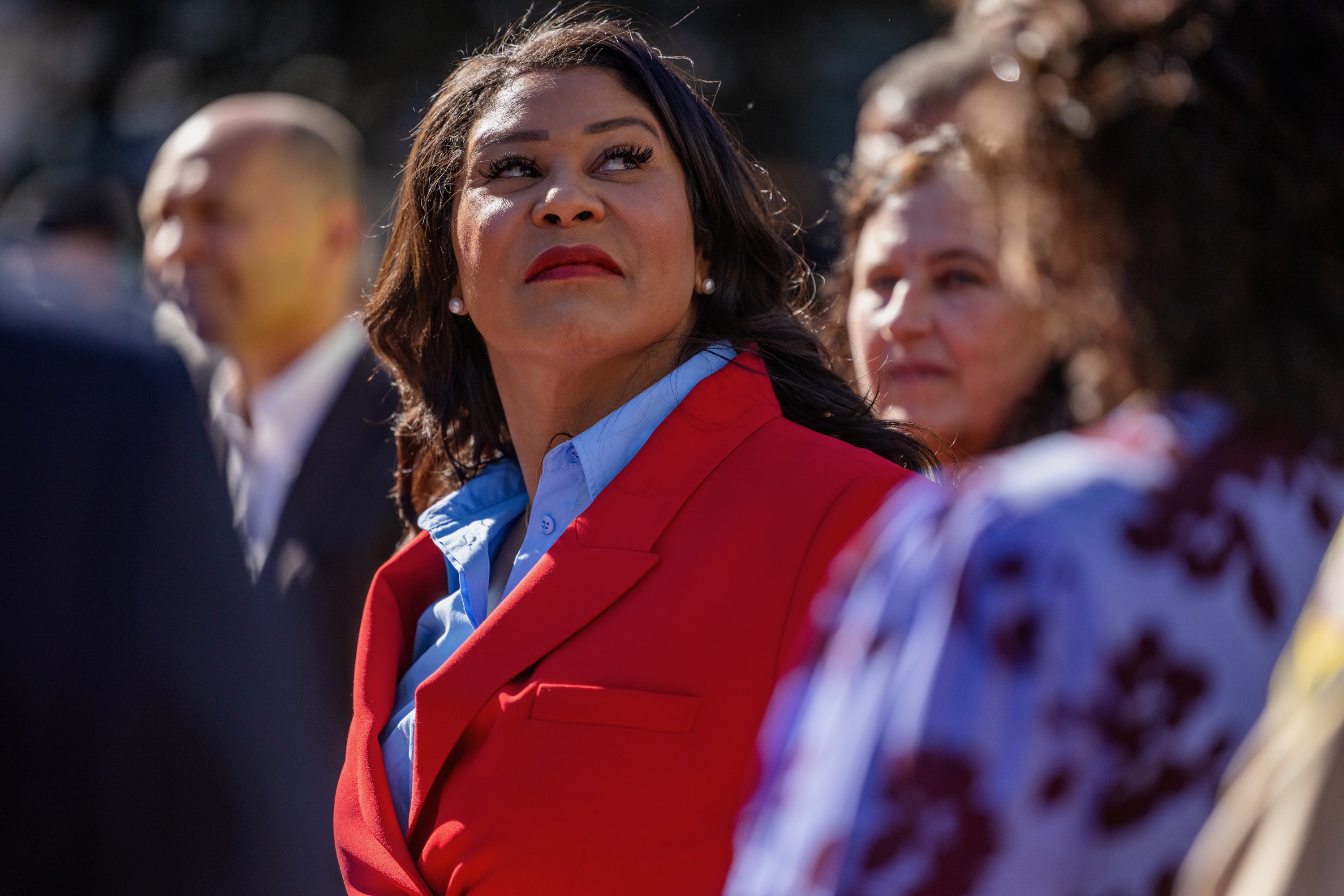 San Francisco Mayor London Breed, with a serious expression, looks towards the sky as she's surrounded by other unidentifiable people that are out of focus.