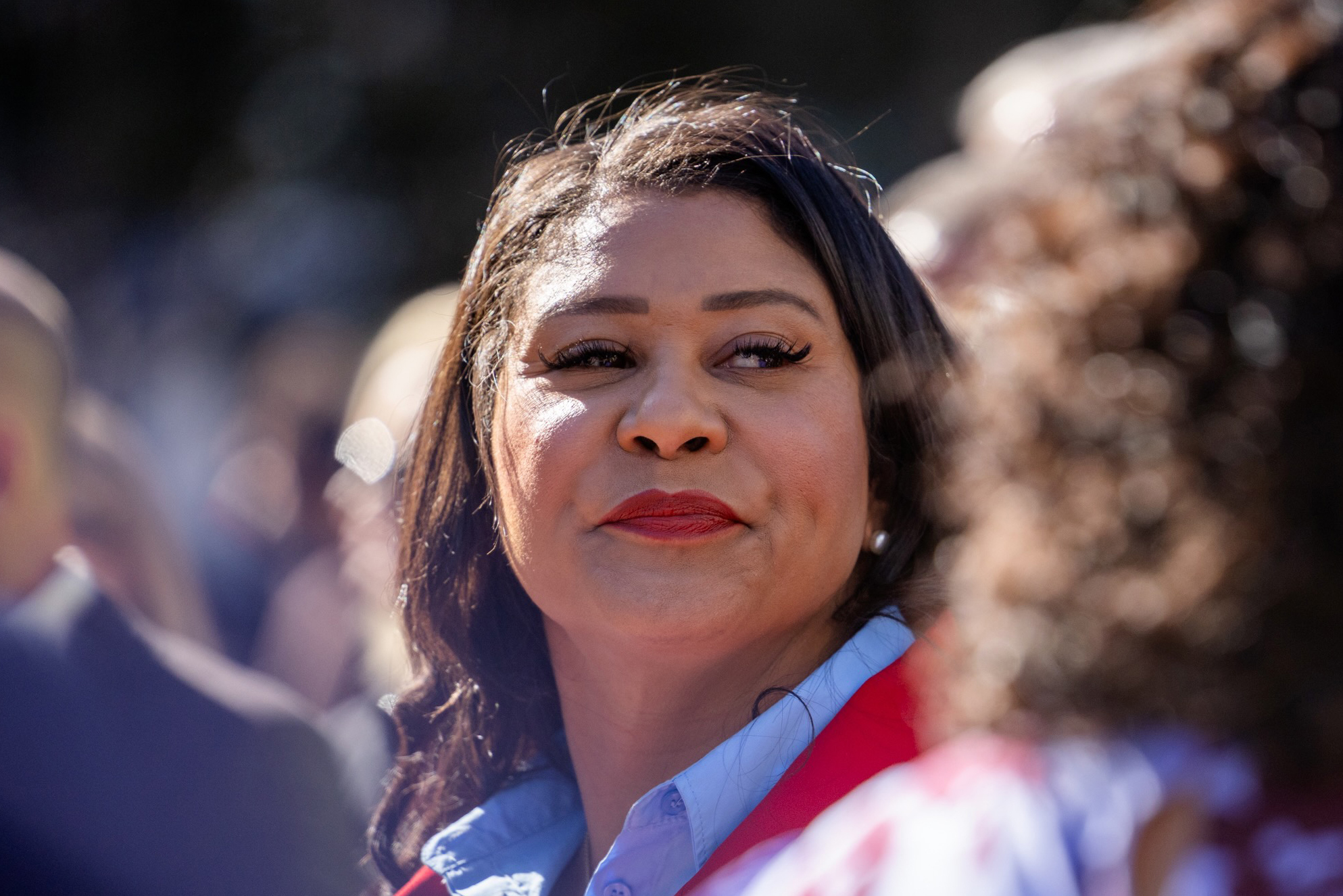 San Francisco Mayor London Breed, with a serious expression, looks towards the right of the frame as she's surrounded by unidentifiable people that are out of focus.