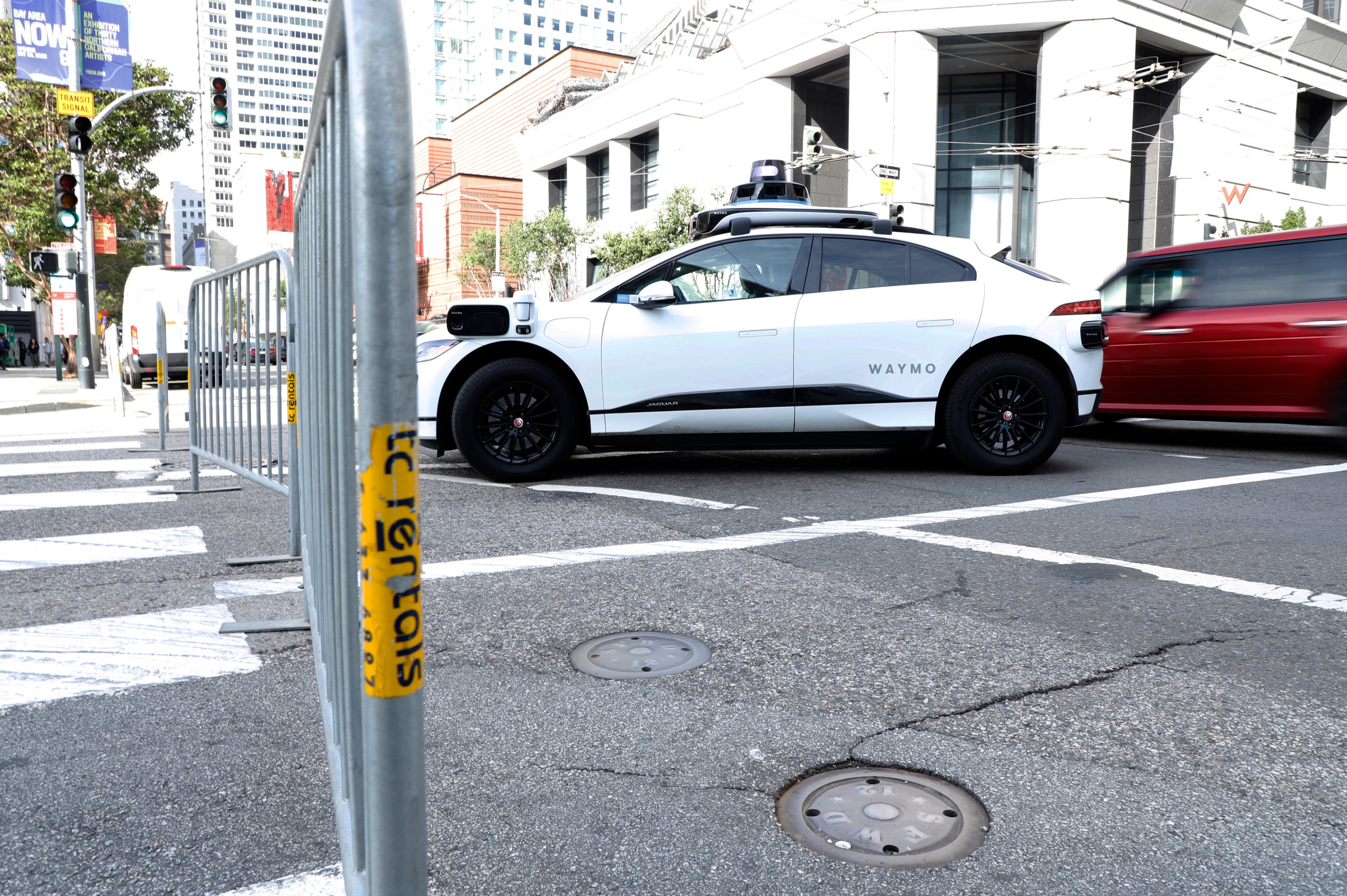 A white Waymo autonomous is idling behind 3 metal barricade on a city street corner as other card pass by.