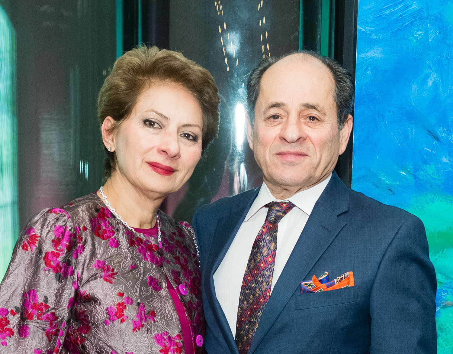 A man dressed in a blue suit and tie stands next to a woman wearing a silver and pink dress as they look at the camera for a posed portrait.