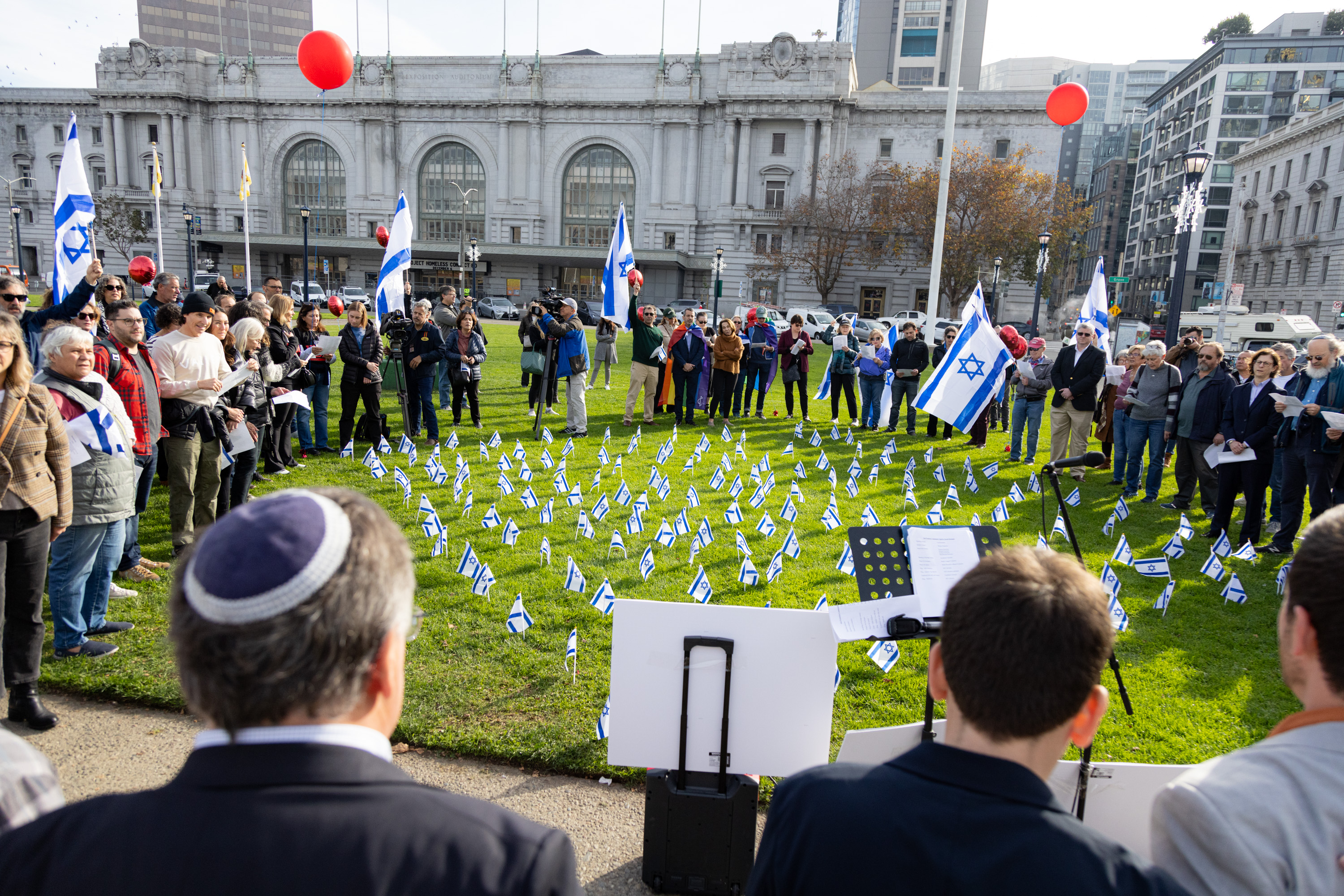 A large group of people gather outside San Francisco City Hall and waves white and blue flags of Israel. Over a hundred small Israeli flags are placed in the ground.