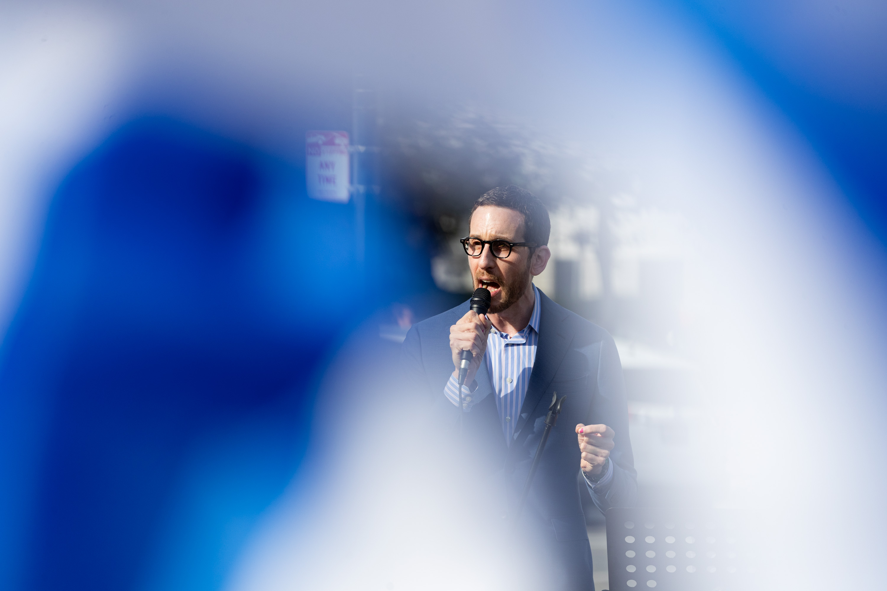 A man in a suit speaks into a microphone, partially obscured by a blue blurry foreground.