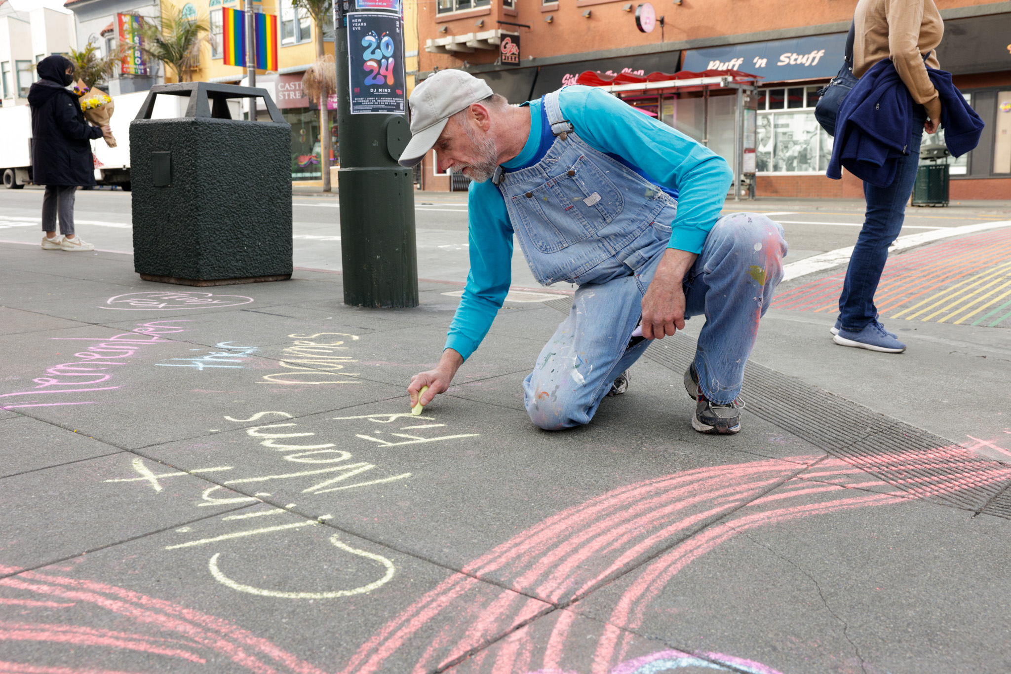 A man wearing a beige-colored baseball cap, a blue long-sleeved shirt and blue denim overalls crouches close to the ground. He has a piece of yellow chalk in his hand and is writing on the ground. A person can be seen behind him in the background to his right, and a telephone poll and trash can are to his left in the background.
