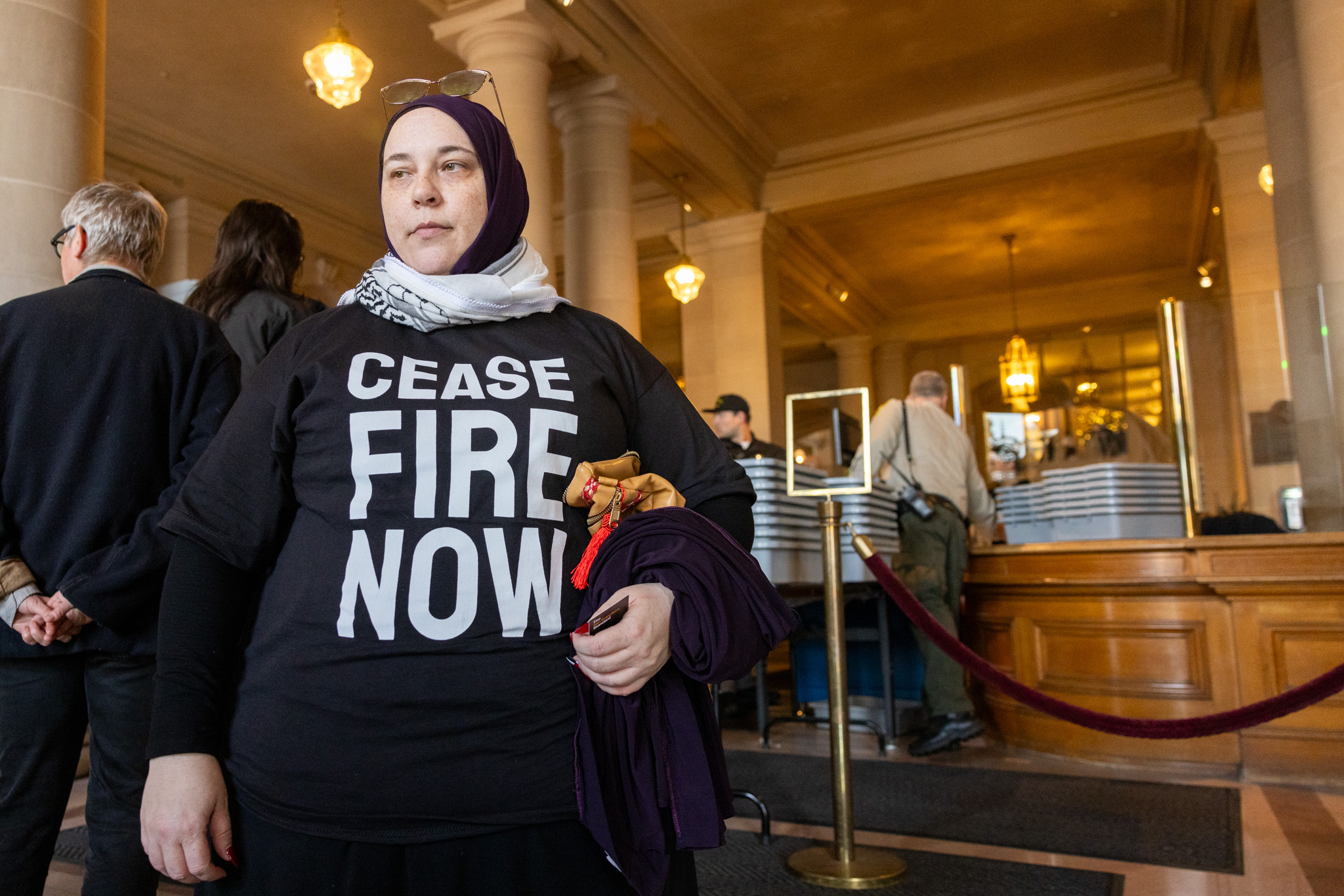A woman wearing a headscarf and a t-shirt reading &quot;Cease-fire now&quot; stands at inside at City Hall.