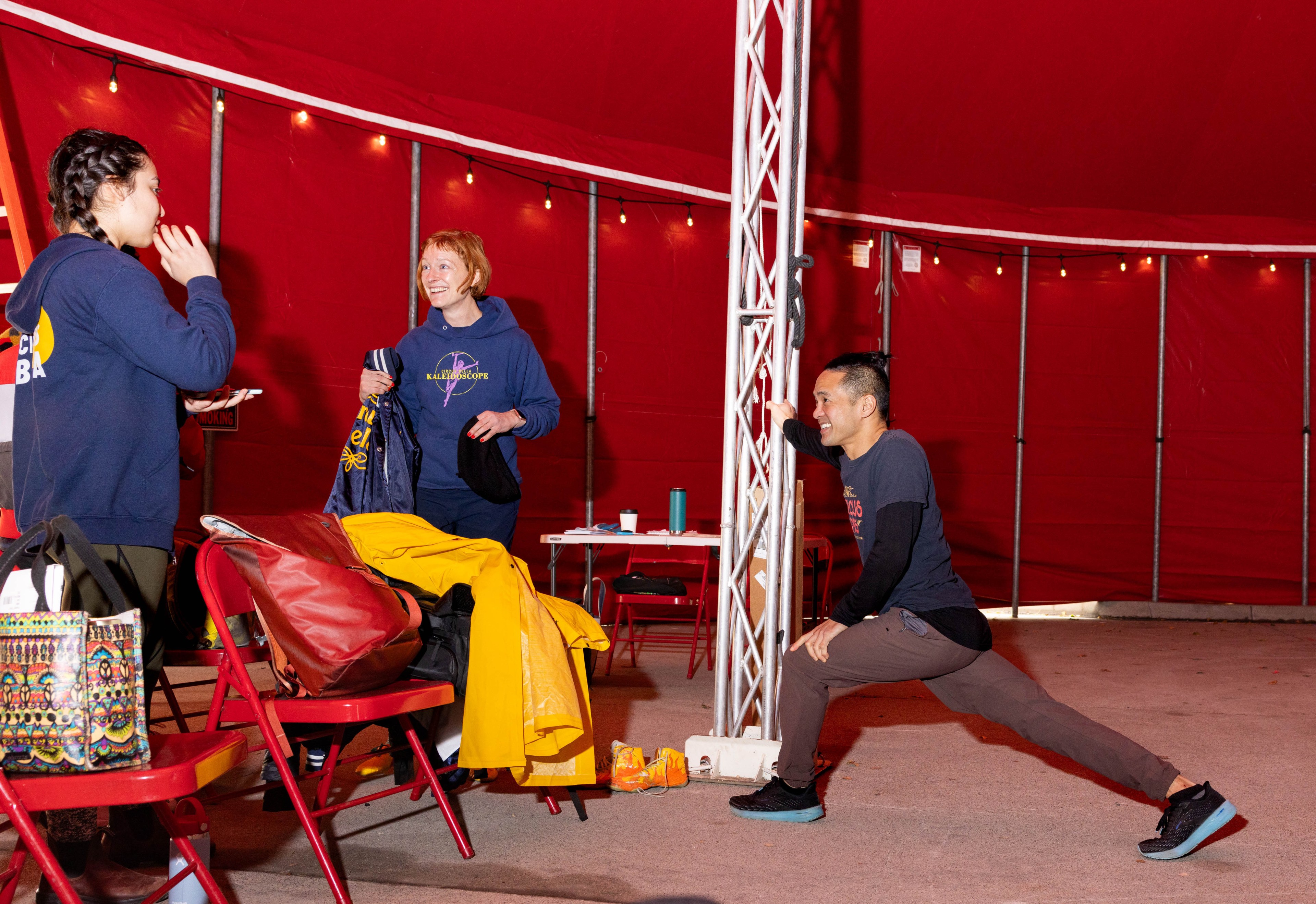 People chat and someone stretches inside the red circus tent.