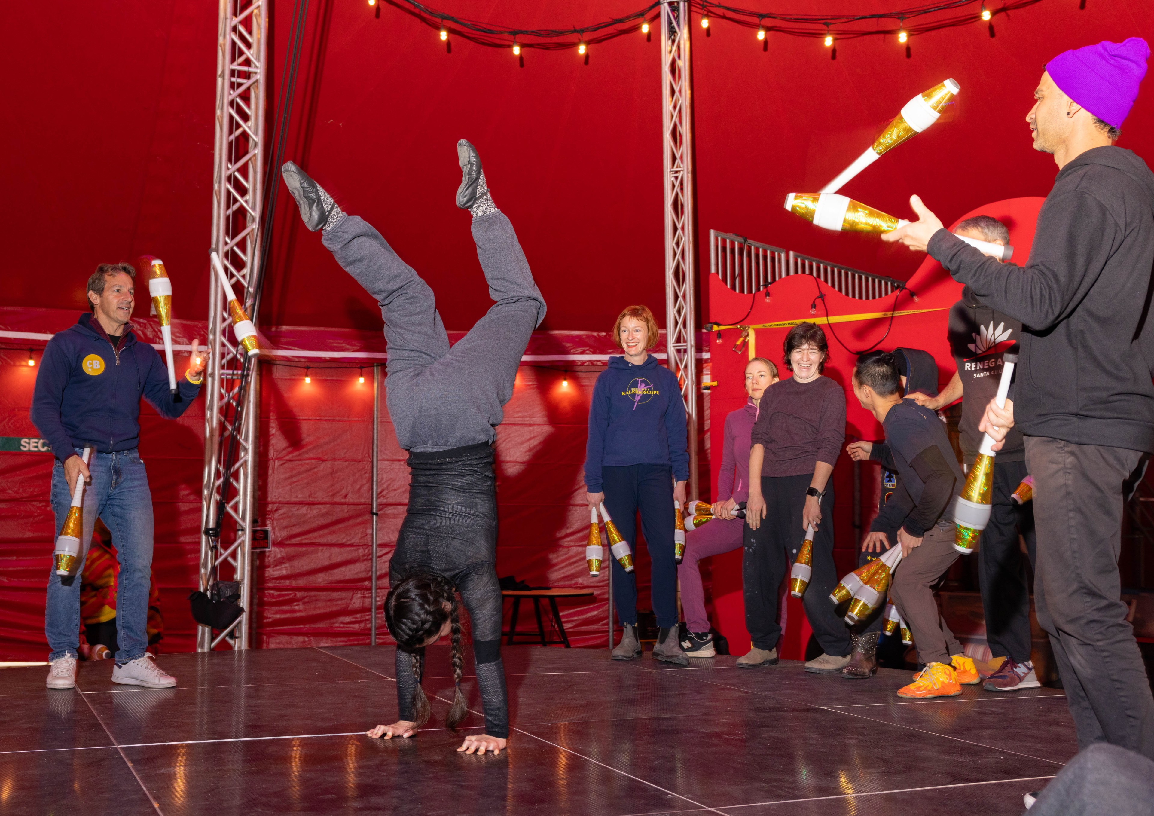 A group of people gather around someone doing a handstand while others juggle.