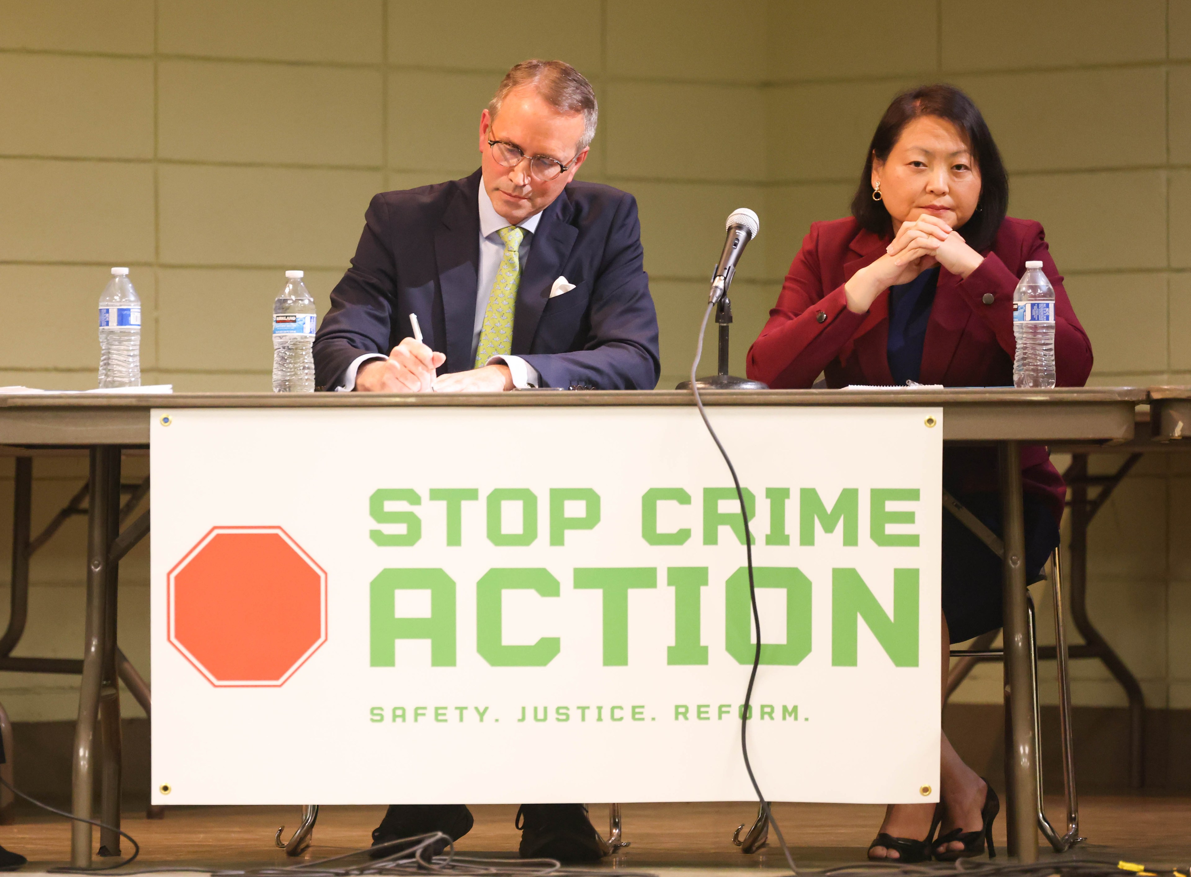 a man in a suit and woman in a red top sit at a table with microphones with a banner that says &quot;stop crime action.&quot;