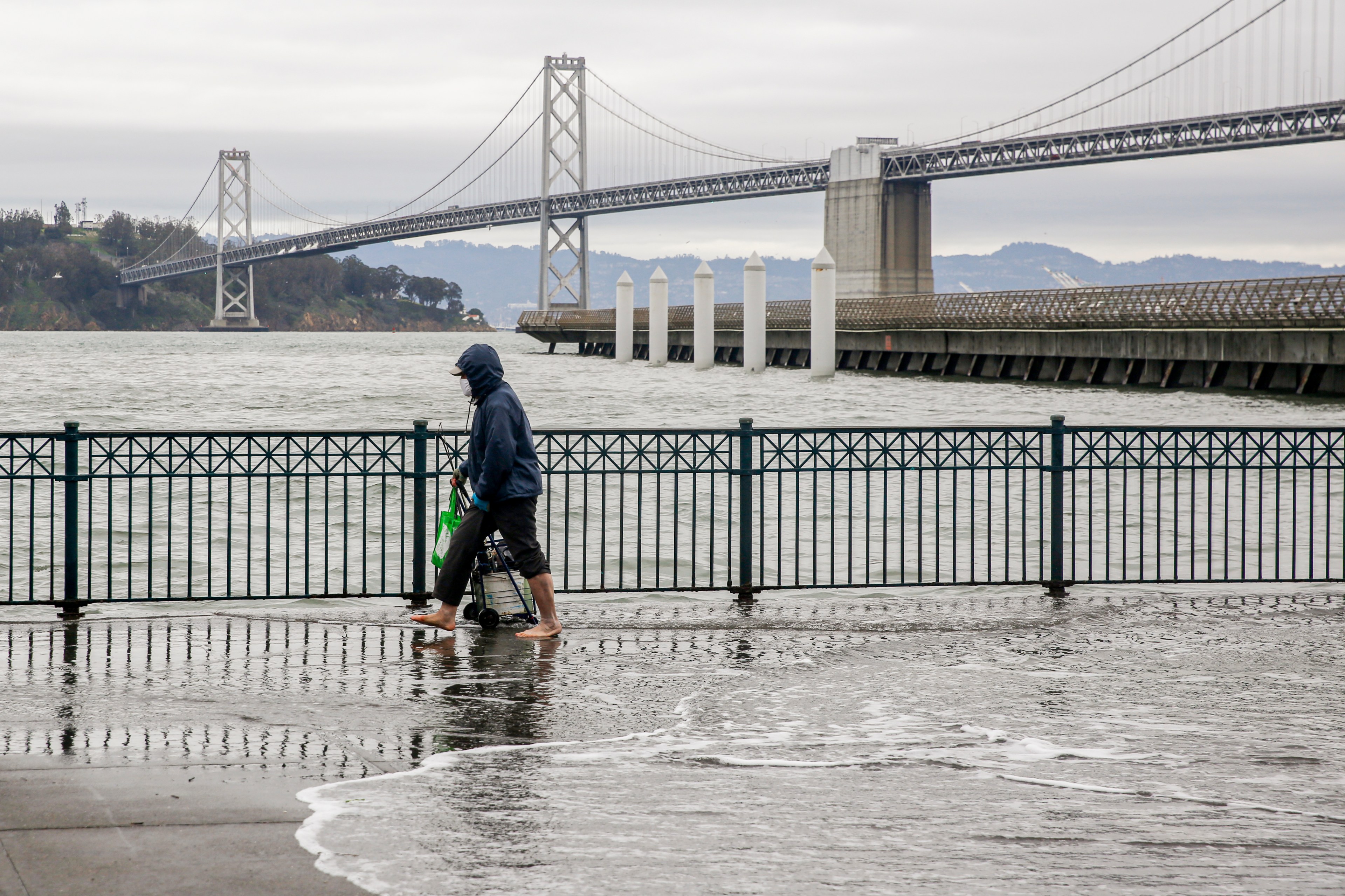 A person walks by flooded bay waters with a bridge in the background.