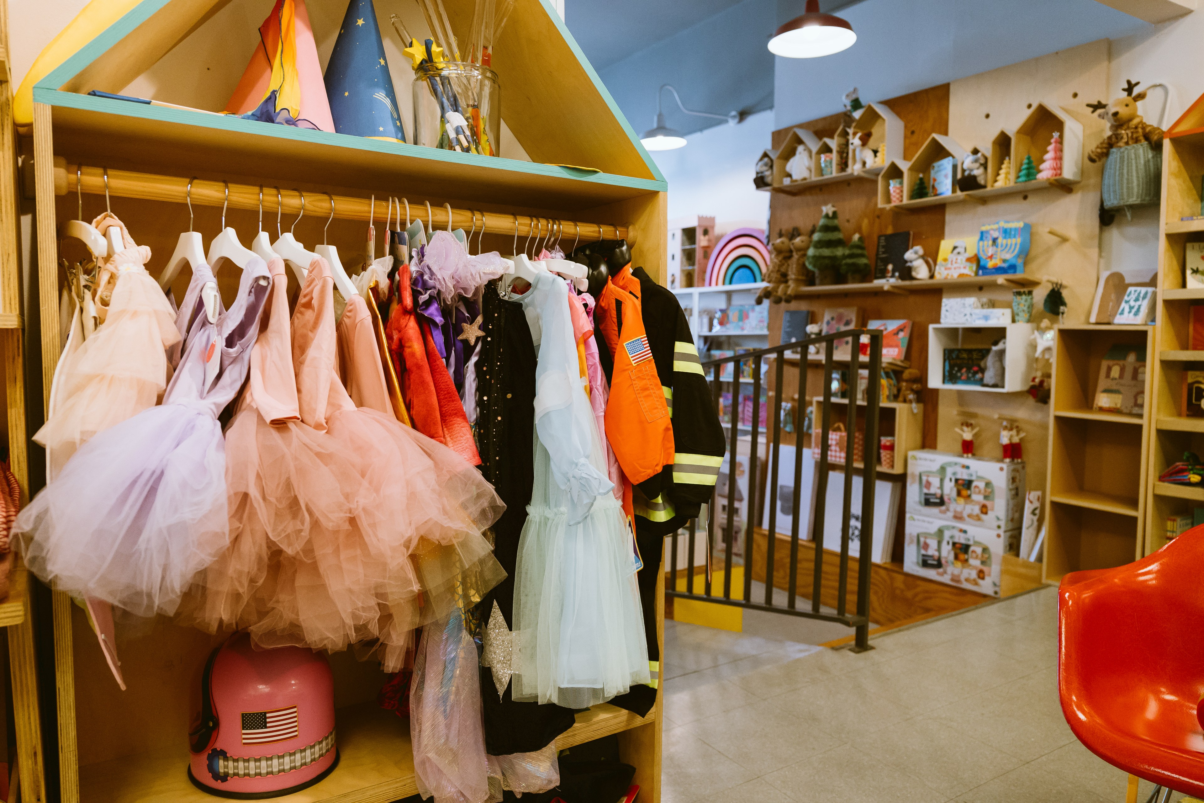 A rack of children's clothing including dresses and tutus with a display of toys in the background.