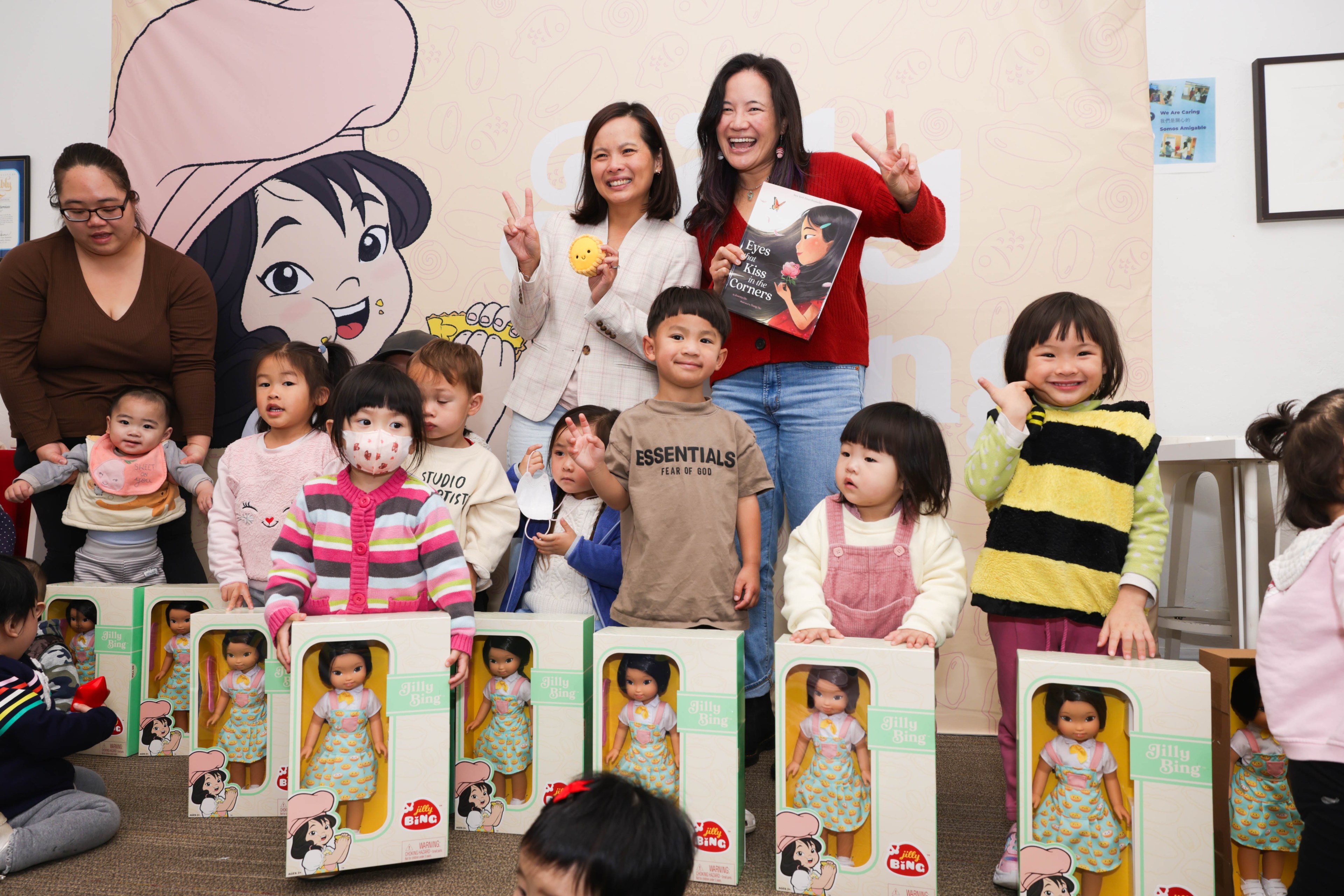 A group of kids and adults in the background pose with dolls in front of them. 