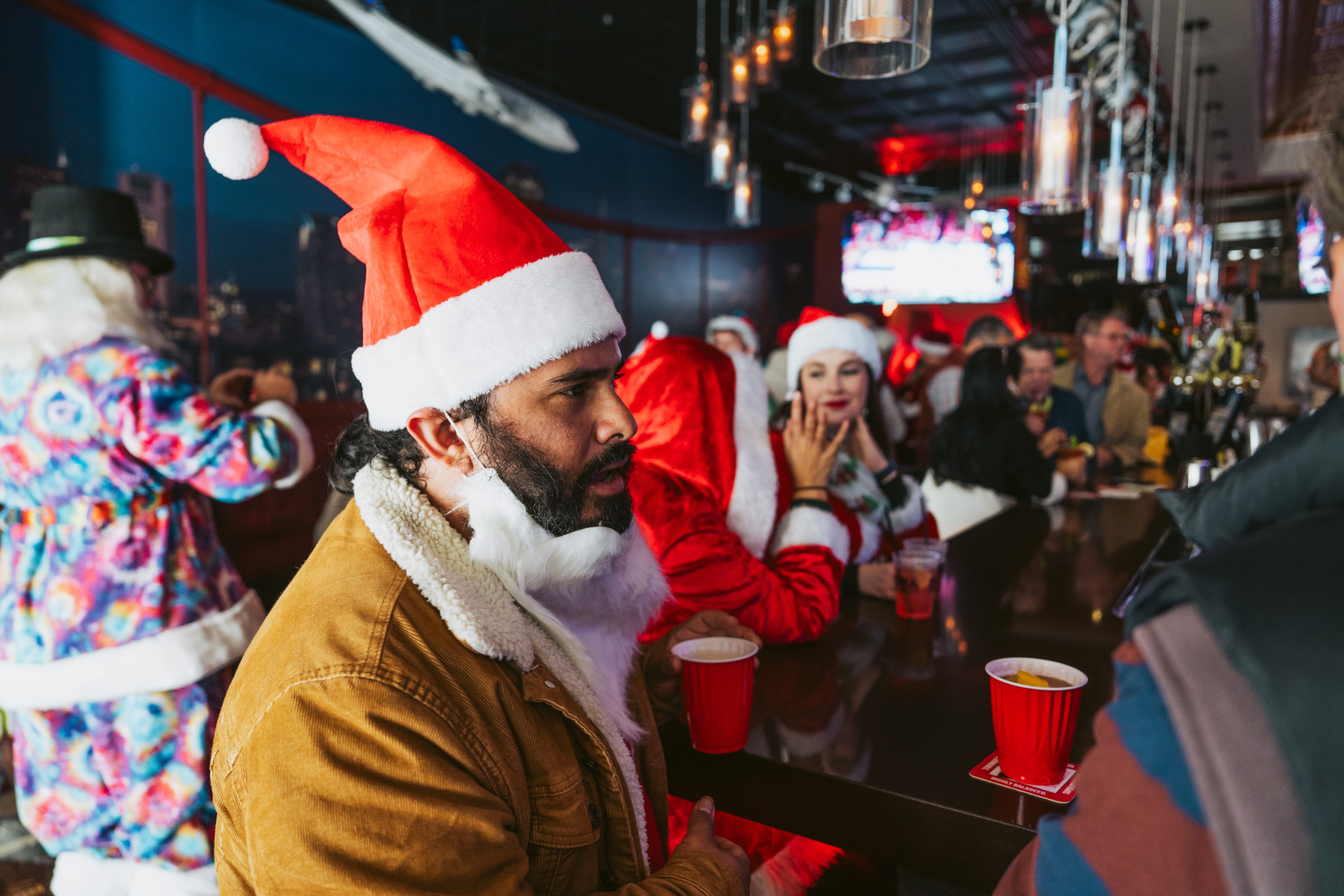 A man in a Santa hat and fake white beard talks to patrons at a bar.