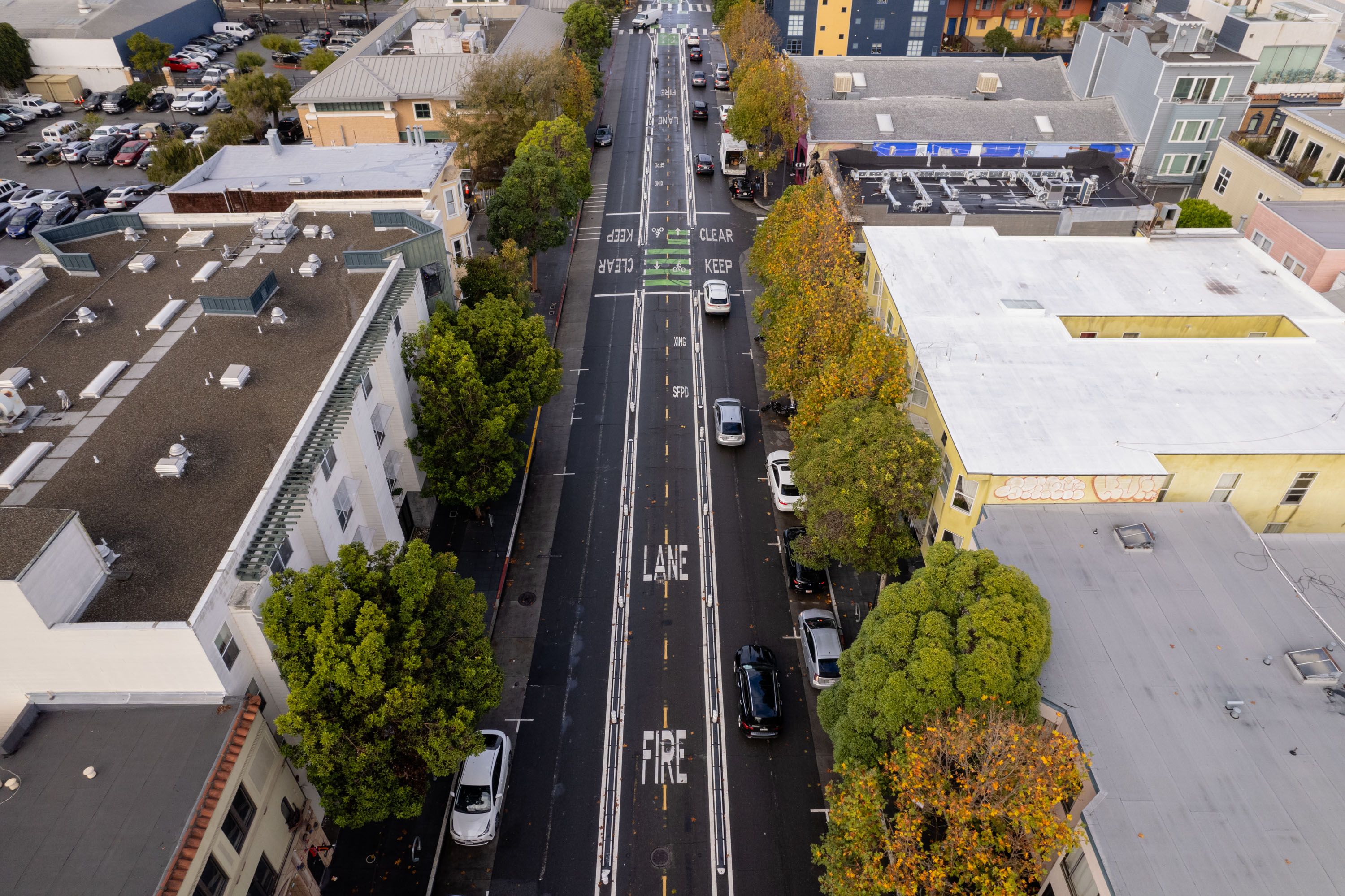 An aerial view of a two-lane road lined with trees and parked cars, flanked by buildings on both sides, with road markings indicating "LANE FIRE," and "KEEP CLEAR."