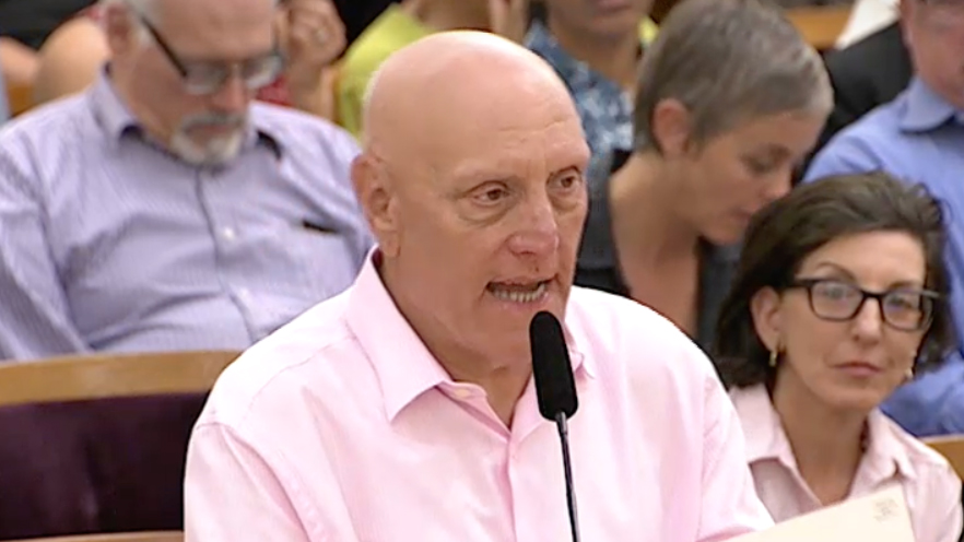 Paul Giusti, a bald man in a pink shirt, speaks into a microphone in a room filled with seated people. Some attendees are paying attention, while others are focused on their own activities.