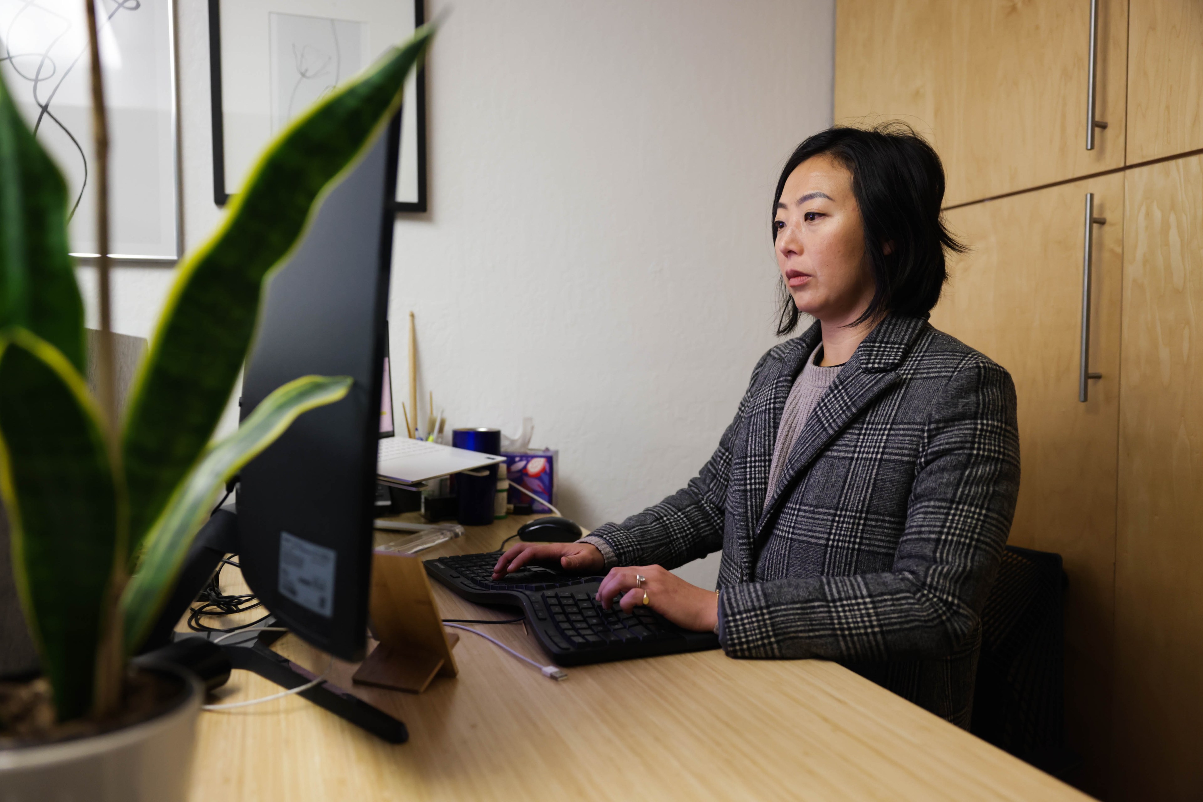 A person at their desk in a gray coat jacket types at a computer.