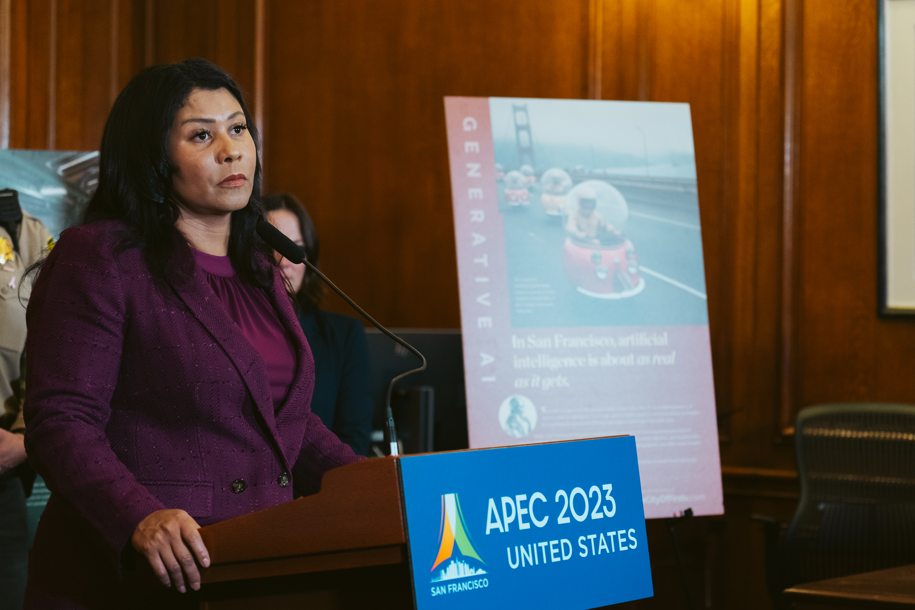 A woman in a magenta jacket speaks at a podium with &quot;APEC 2023 UNITED STATES&quot; displayed, with a poster about AI and San Francisco's Golden Gate Bridge in the background.