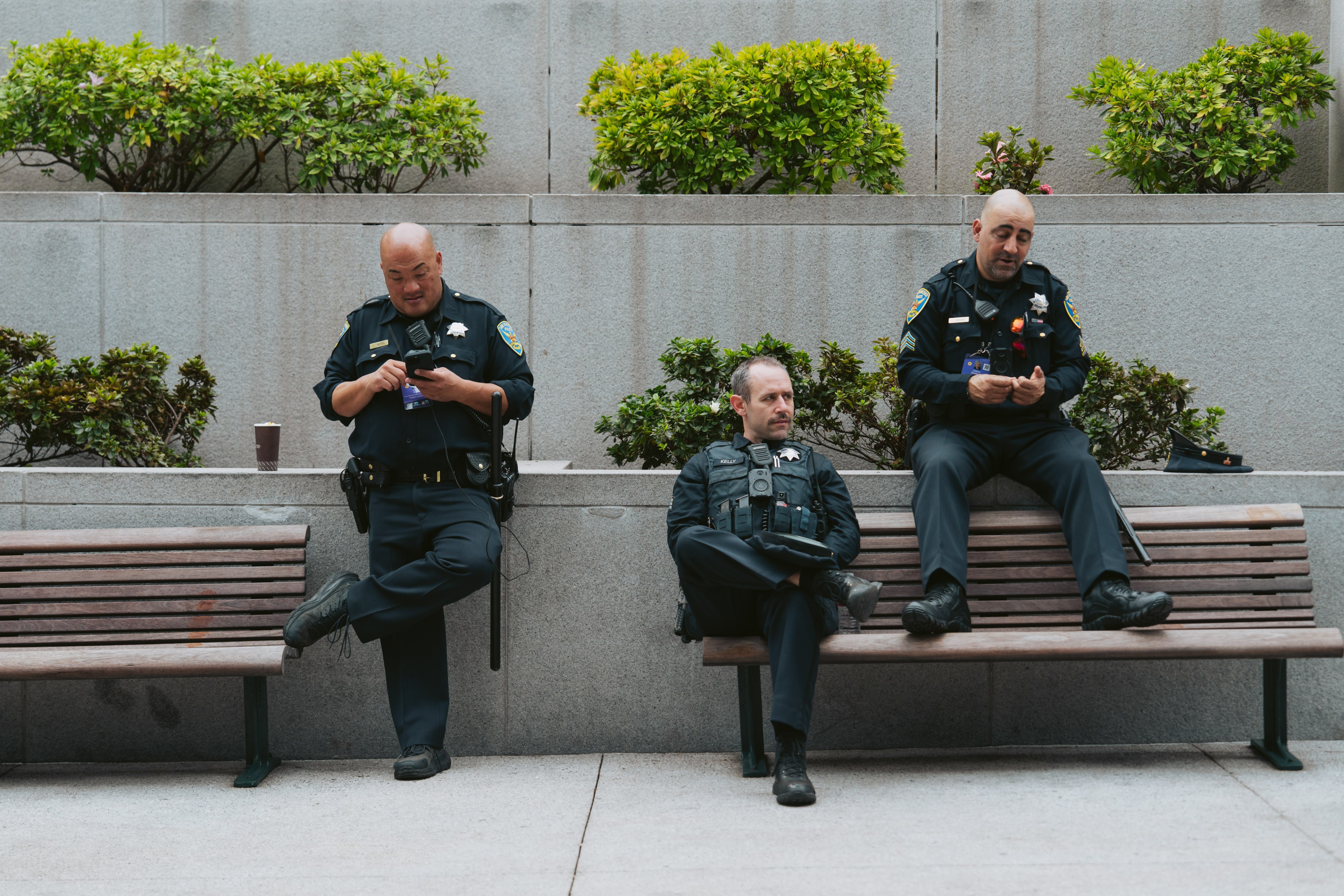 Three police officers are sitting on a bench; two are on their phones, and one looks pensive. There's greenery behind them on a gray wall.