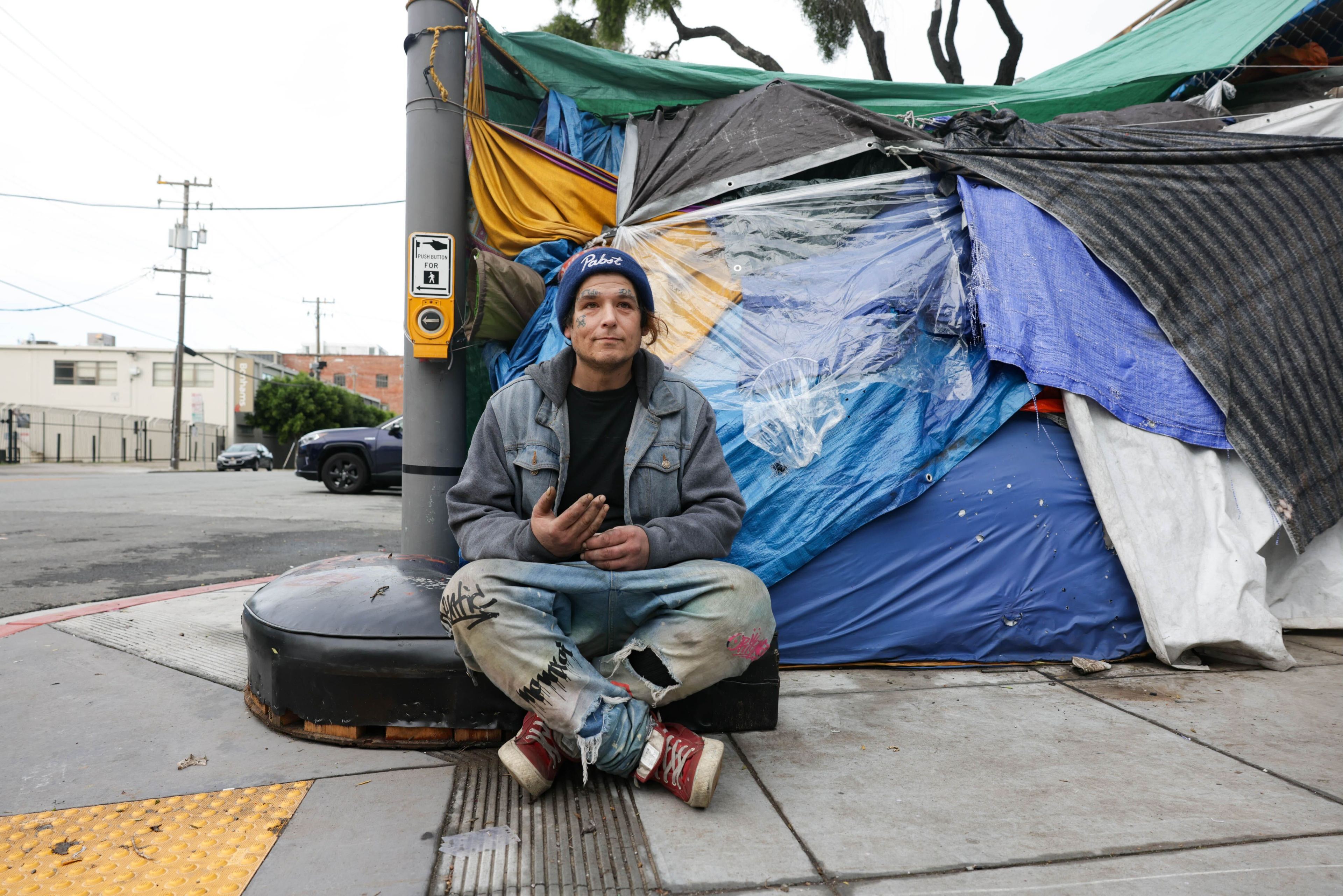 A man sits on a sidewalk near a makeshift tent, suggesting urban homelessness.