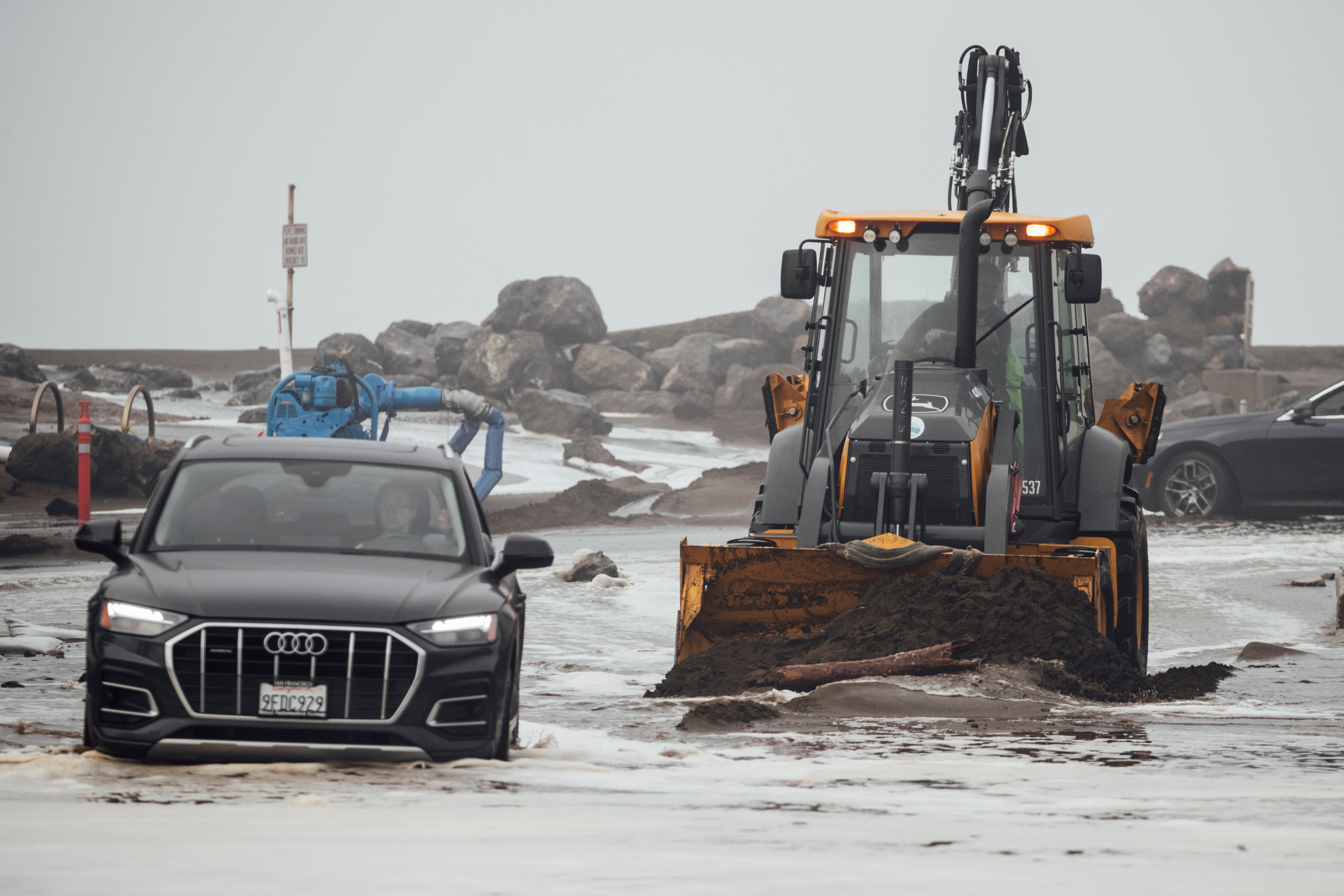 A dark colored sedan sits in surging water beside a bulldozer on a cloudy day near a rocky oceanfront shoreline.
