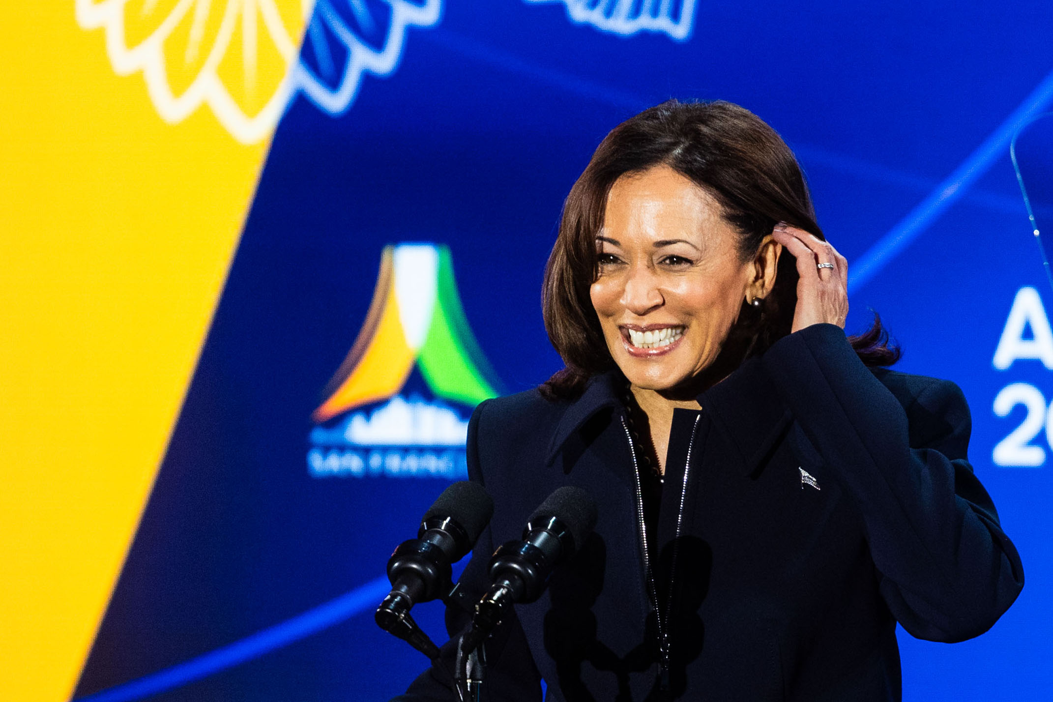A woman smiles, touching her ear, standing at a podium with microphones, against a vibrant logo backdrop.