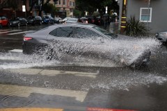A car drives through a puddle, creating a large splash on a wet urban street.
