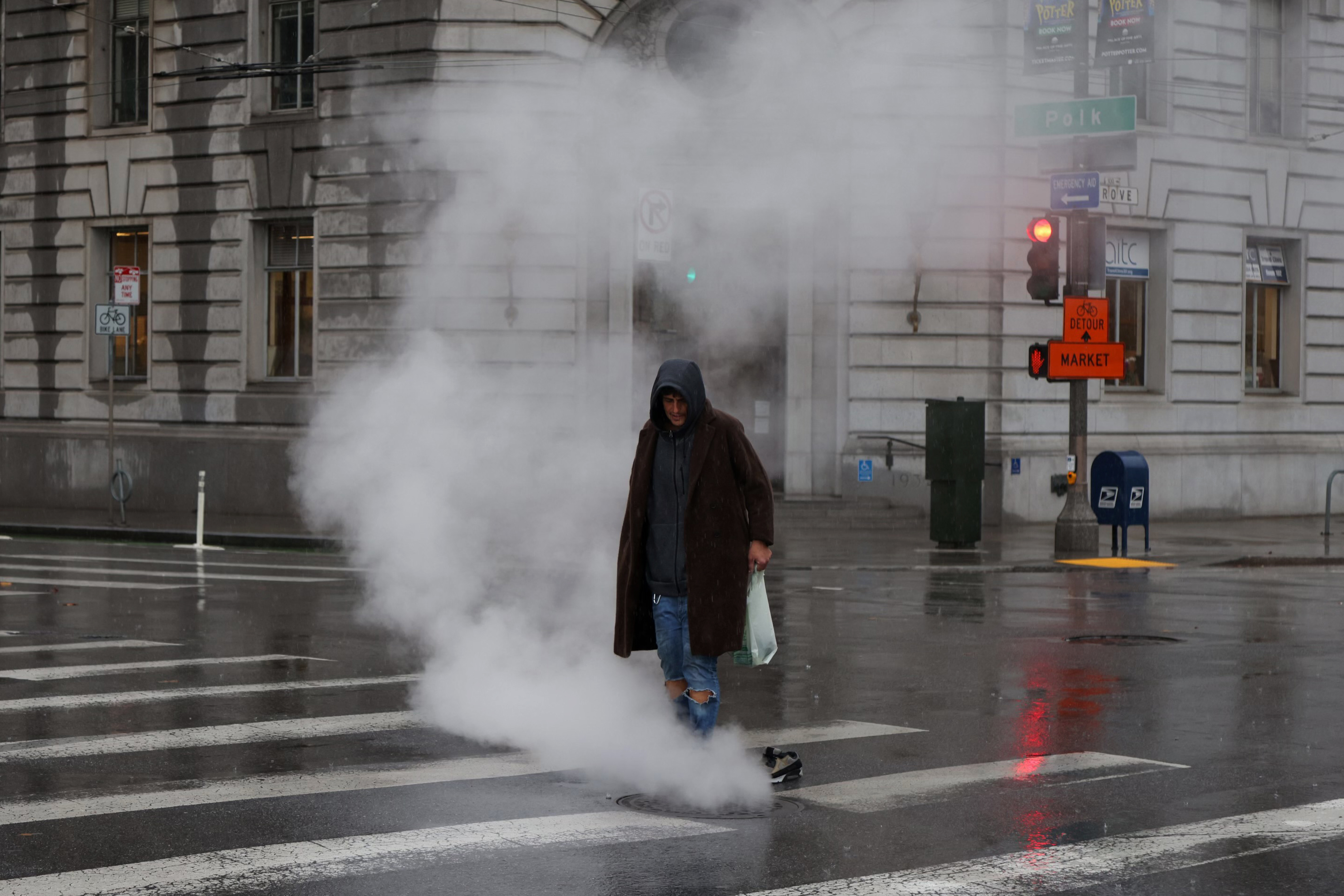 A person crosses a wet street with steam rising from a manhole. It's a gloomy, overcast day.