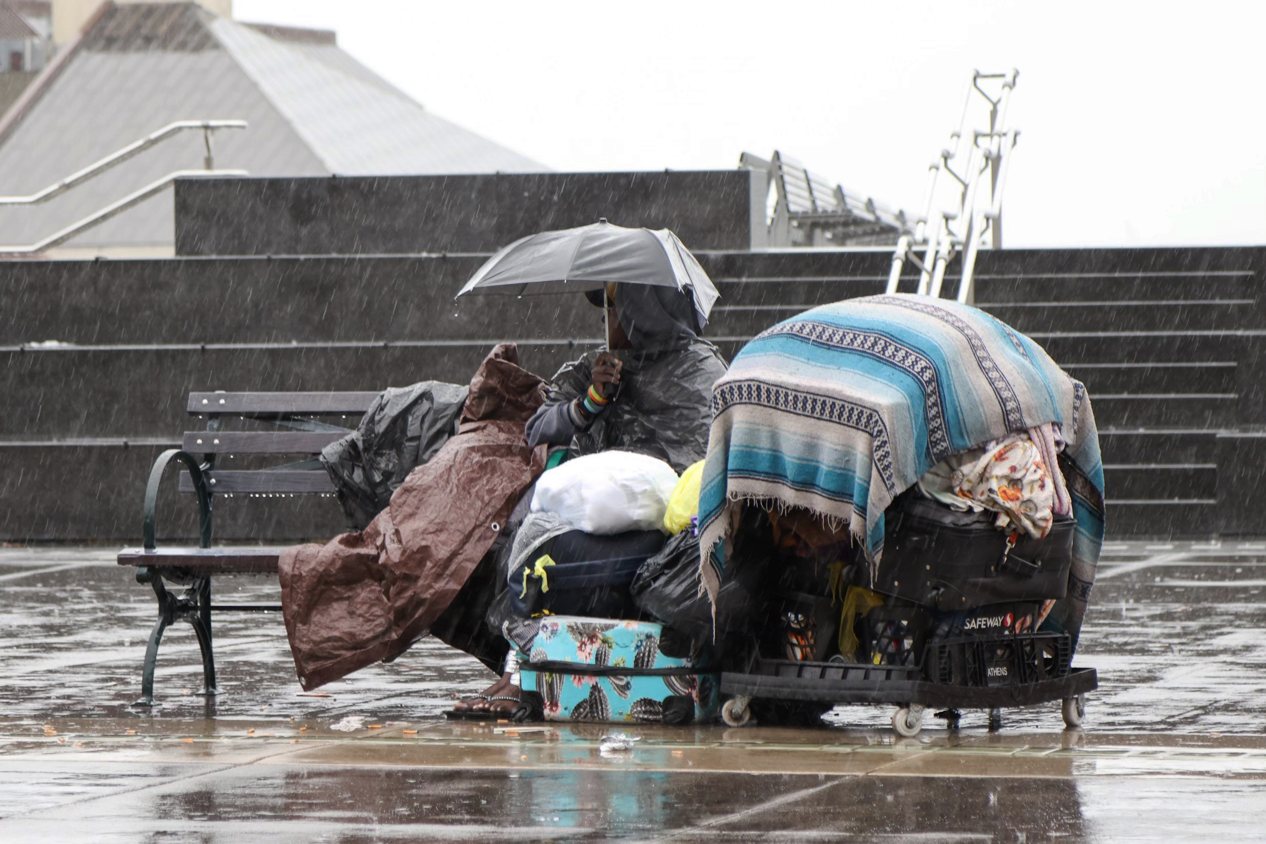 A person under an umbrella sits on a bench in the rain, surrounded by belongings covered with a tarp.
