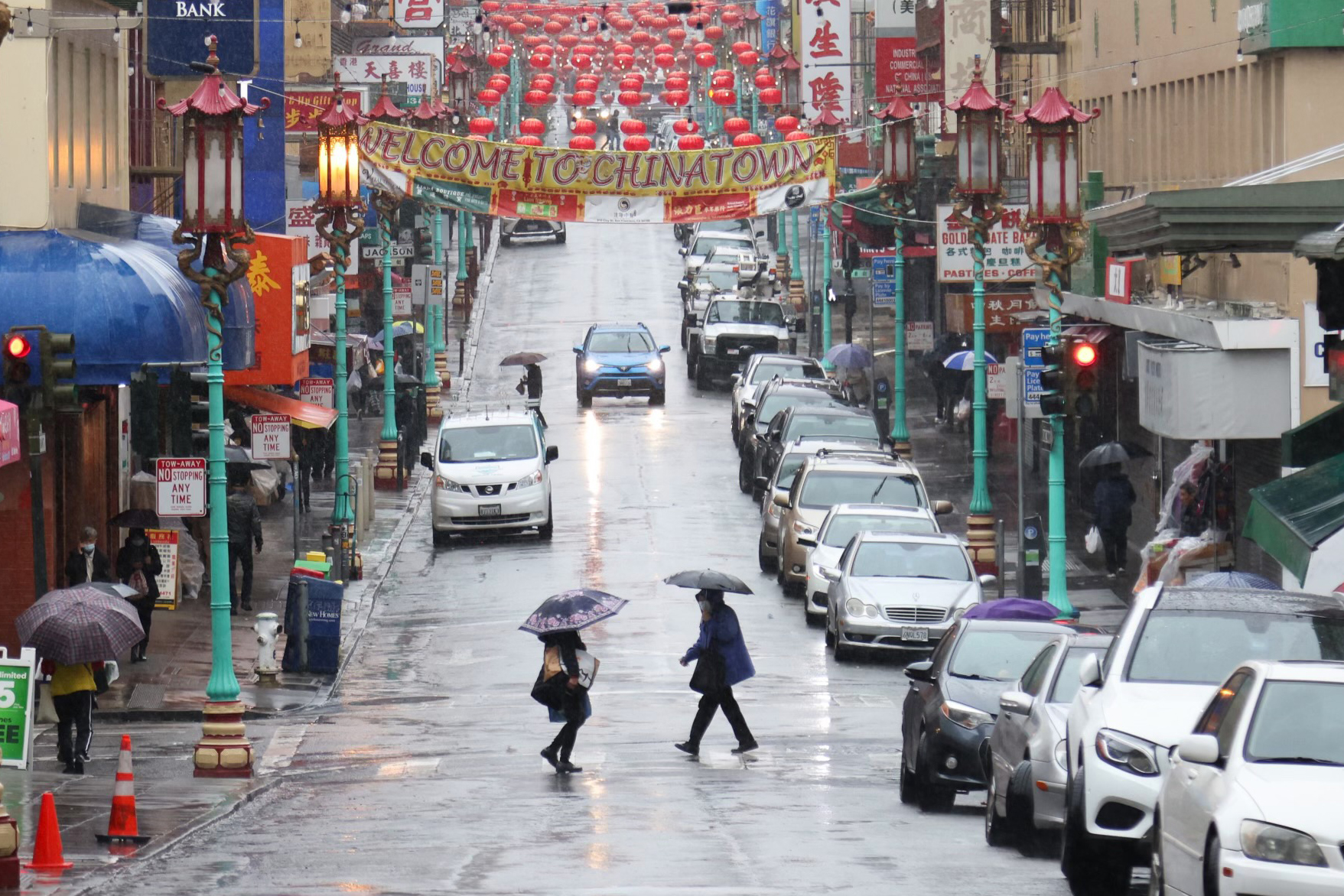 A rainy street in Chinatown with pedestrians holding umbrellas, lined with parked cars and red lanterns overhead.