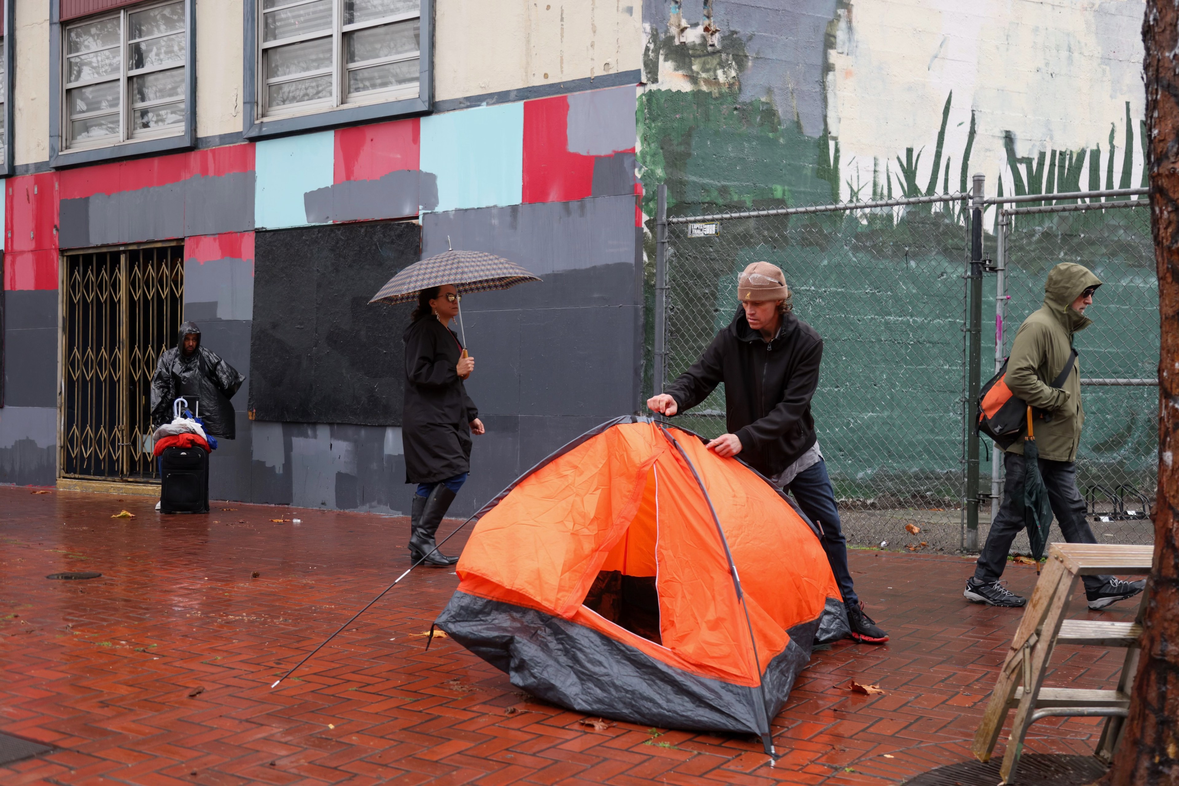 A person sets up an orange tent on a wet city sidewalk, while others with umbrellas pass by a colorful mural wall.