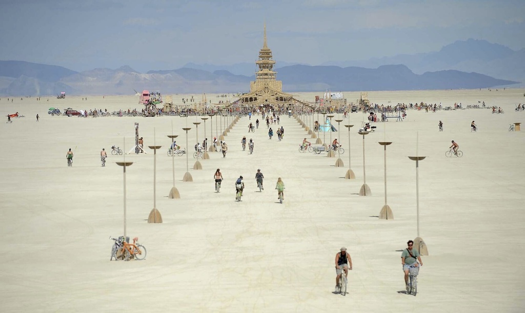 People cycle on a flat plain towards a large, ornate wooden structure, with lamp posts lining the path under a clear sky.