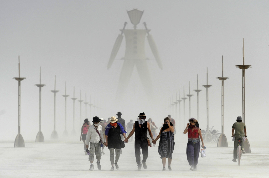 A group of people walks across a dusty, flat expanse towards a large figure, with rows of poles receding into the haze.