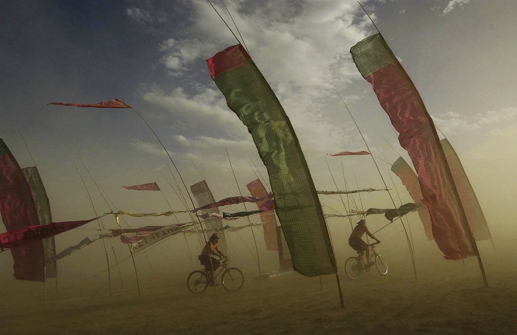 Two people biking among tall, colorful flags in a hazy, dust-filled environment.