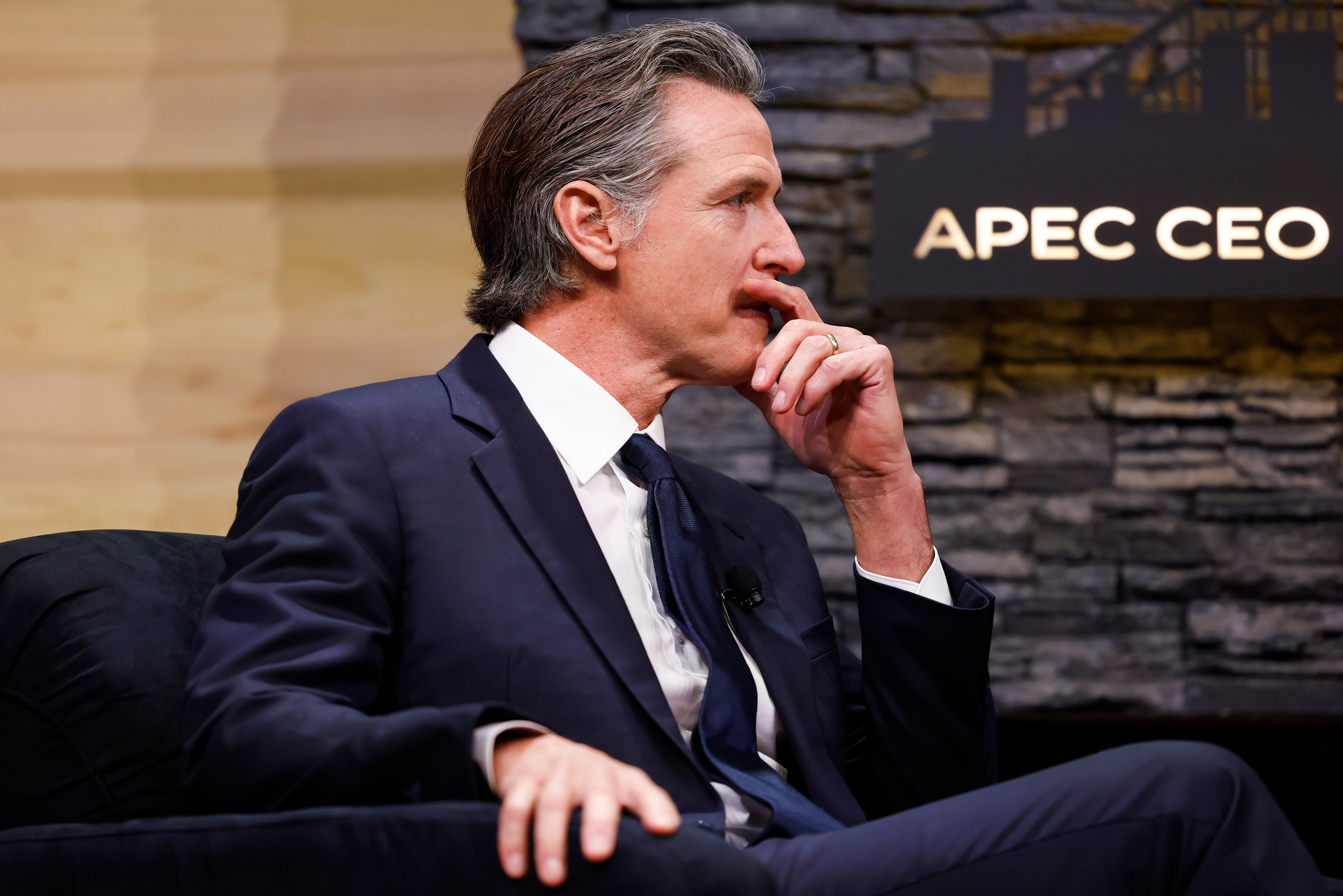 A man, Governor Gavin Newsom, sits in a chair, looking to the right with his hand on his chin as if in thought.